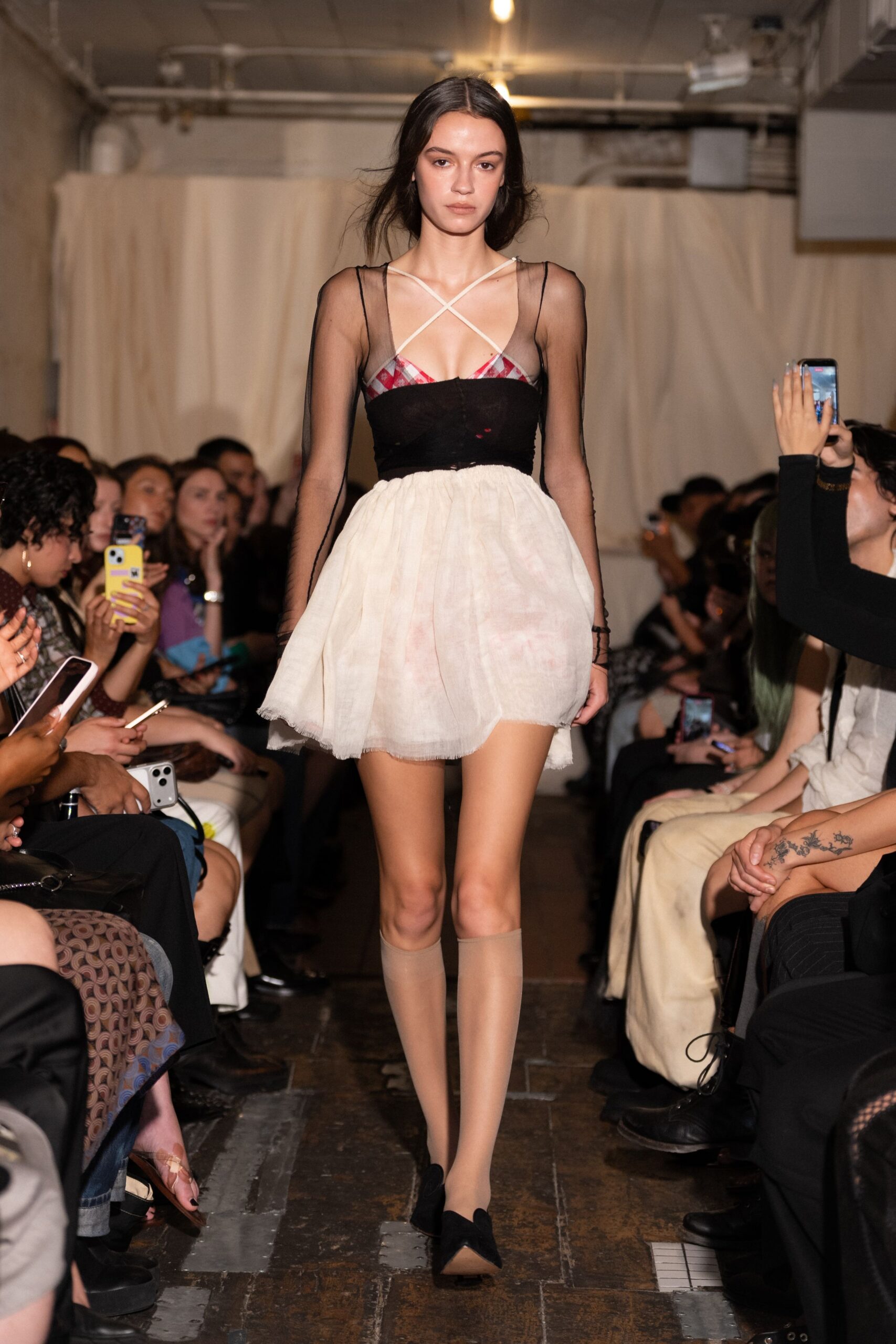 Sheer black mesh bodice over a plaid bra top, crossed ivory straps. Bubble mini skirt in gauzy cream, faintly blushed and raw at the hem. Nude knee socks, slightly darker band at the top. Black pointed flats with shallow vamp. Walking into a church basement recital with your cardigan left behind.