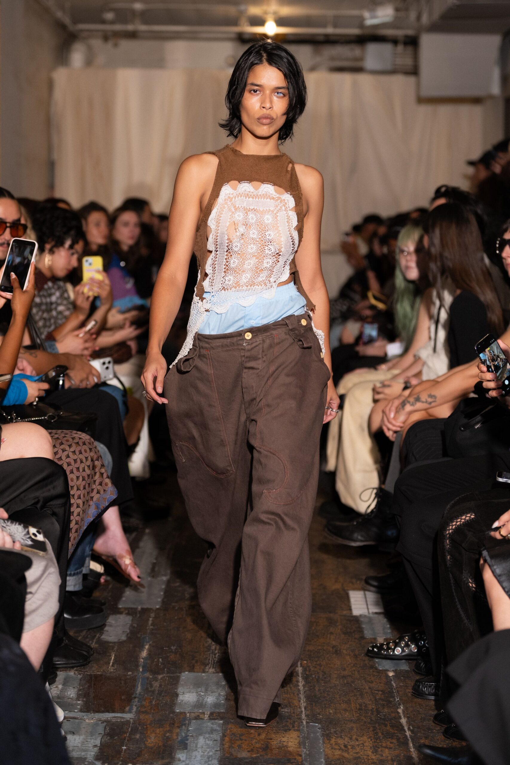 Brown open-knit tank shredded around a white lace front panel. Powder-blue shirting peeks out in a soft bloused band. Loose dark-brown work trousers, offset buttons and curved knee patches. Square-toe shoes barely visible under the hem. For crossing the laundromat floor with your shirt half tucked back in.