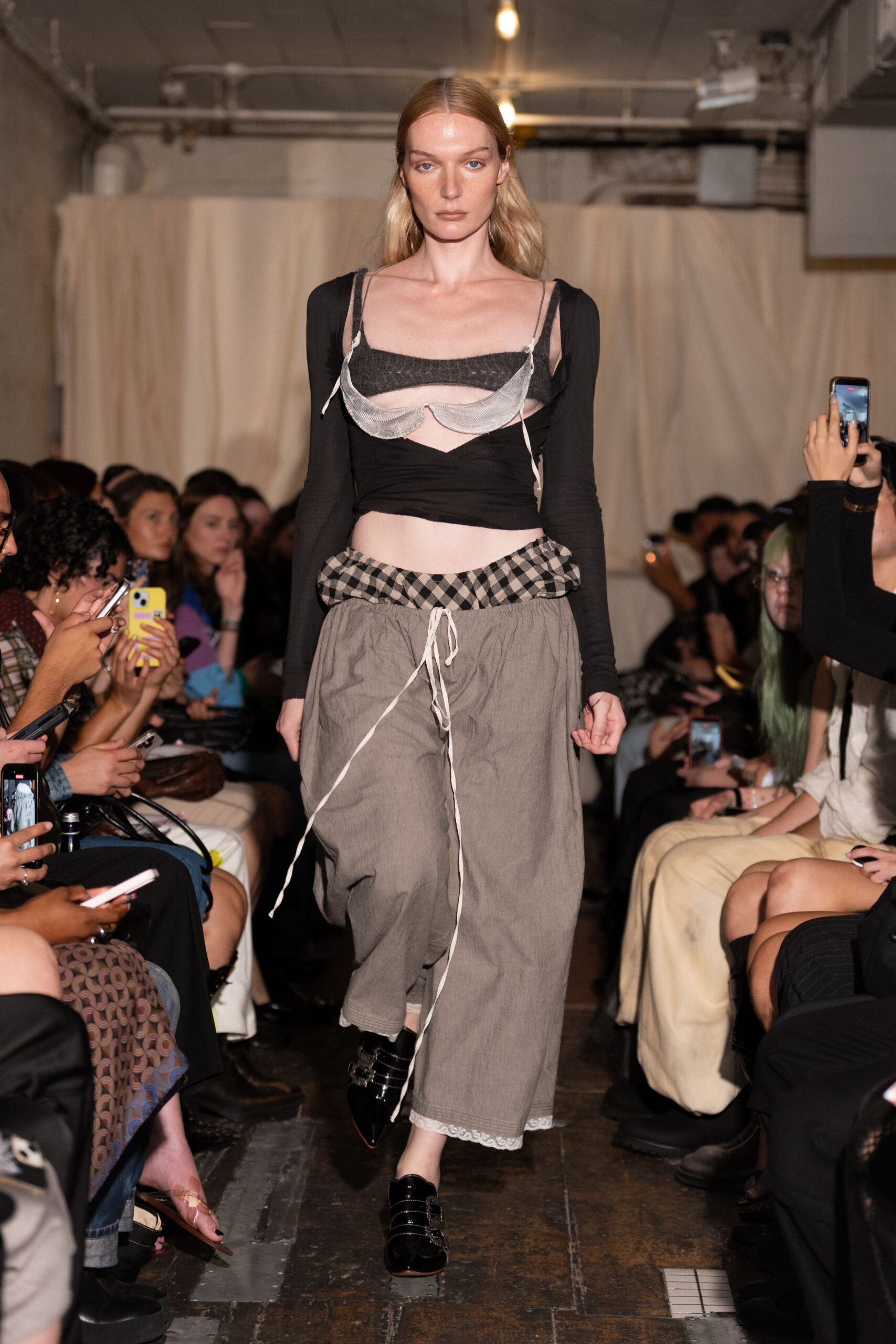 Sheer white bra slung over the chest, ties hanging loose. Black jersey crop top with a cutaway bust band. Dusty grey wide trousers, gingham waistband puffed above a long drawcord. Black patent monk shoes with stacked buckle straps. Whatever you pull on for dawn coffee after sleeping in borrowed clothes.