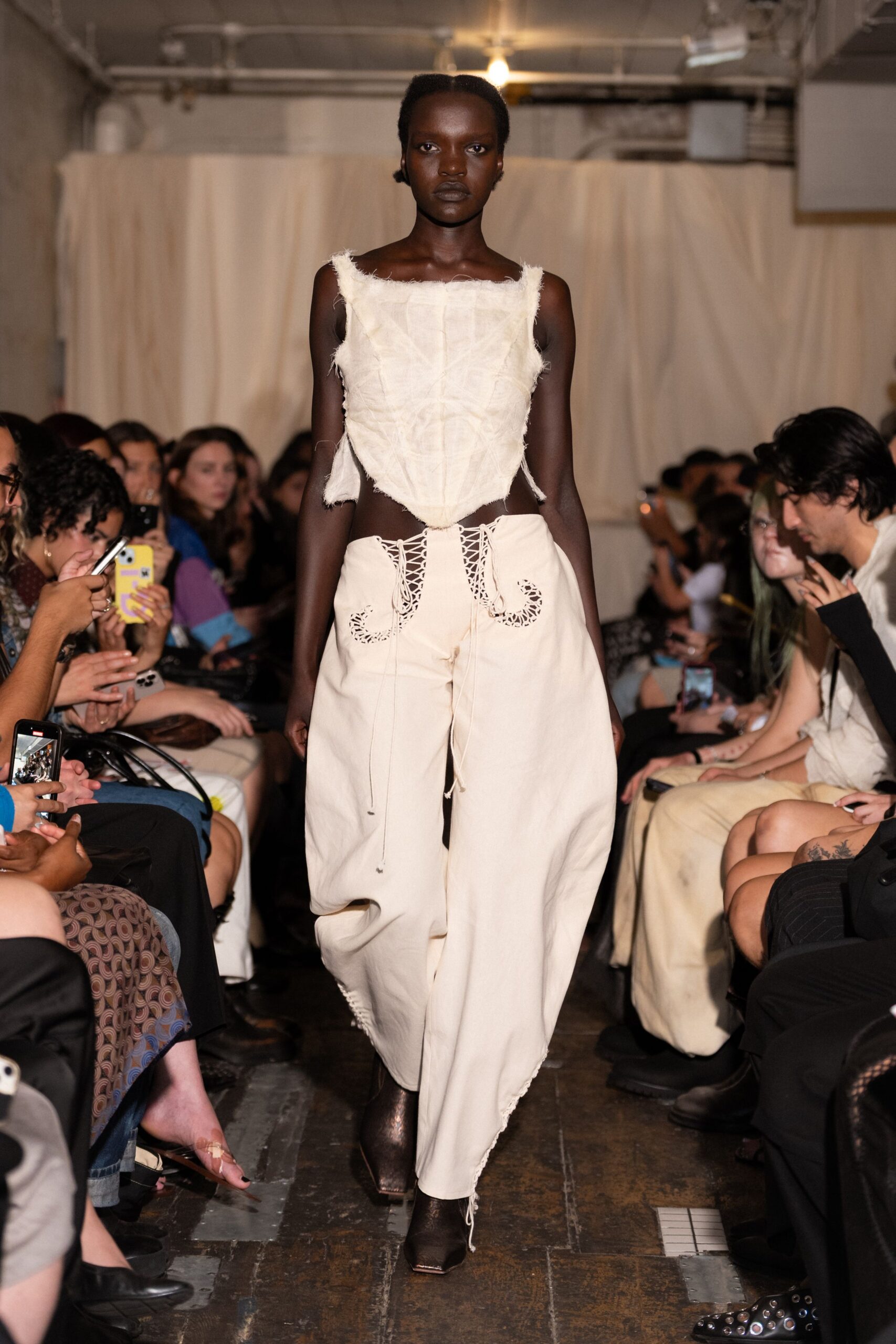 Raw-edged ivory felted top, seams sketched in darker thread. Ballooning cream trousers with corset-laced hips and cutout pocket scrolls. Dark square-toe boots peeking under the dragging hem cords. For crossing a drafty studio floor with plaster dust still in the air.