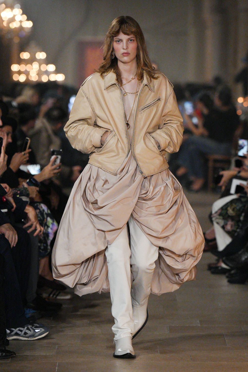 Buttercream leather bomber with dropped shoulders and a slanted chest zip. Blush satin dress scooped low, gathered into heavy pouf swags. Long gold chain with mismatched pendants. White pointed boots, creased hard across the instep. Whatever you wear home after sleeping through checkout at the old hotel.