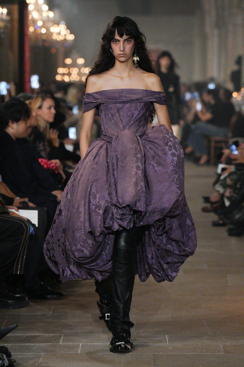 Off-shoulder plum brocade gown, corseted through the torso and gathered into swollen pannier folds. Black leather trousers disappearing beneath the lifted skirt. Slouching black boots with square silver buckle straps. Mismatched drop earrings. Crossing a drafty manor corridor after everyone else has gone to bed.