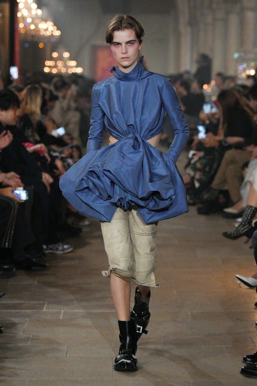 Steel-blue taffeta top with a high neck and twisted front knot. Matching taffeta bubble skirt, ballooned into a dense windblown pouf. Bone leather cropped trousers, creased and strapped loose below the knee. Black ankle boots with broad silver buckles. Crossing a drafty palace corridor after knocking over a centerpiece.