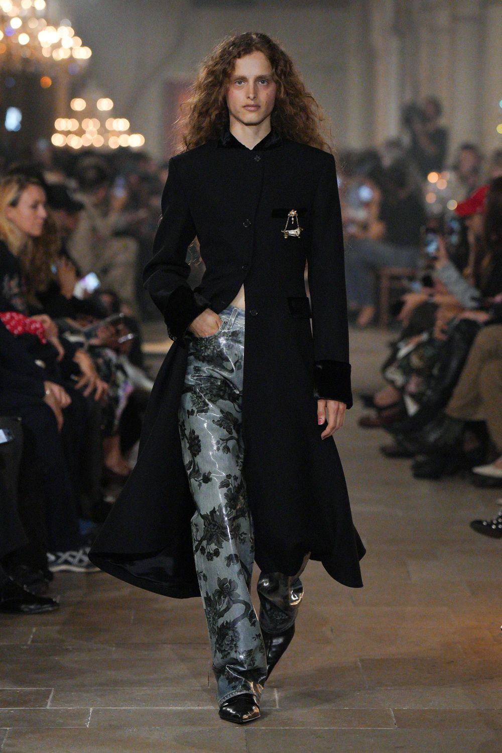 Long black tailored coat, velvet pocket welts and deep turned cuffs. Tiny silver rocking-horse brooch pinned at the chest. Pale blue satin trousers overprinted with inky black florals. Black pointed boots with a hard metallic toe cap. Crossing a drafty manor hall after midnight, still carrying the party with you.