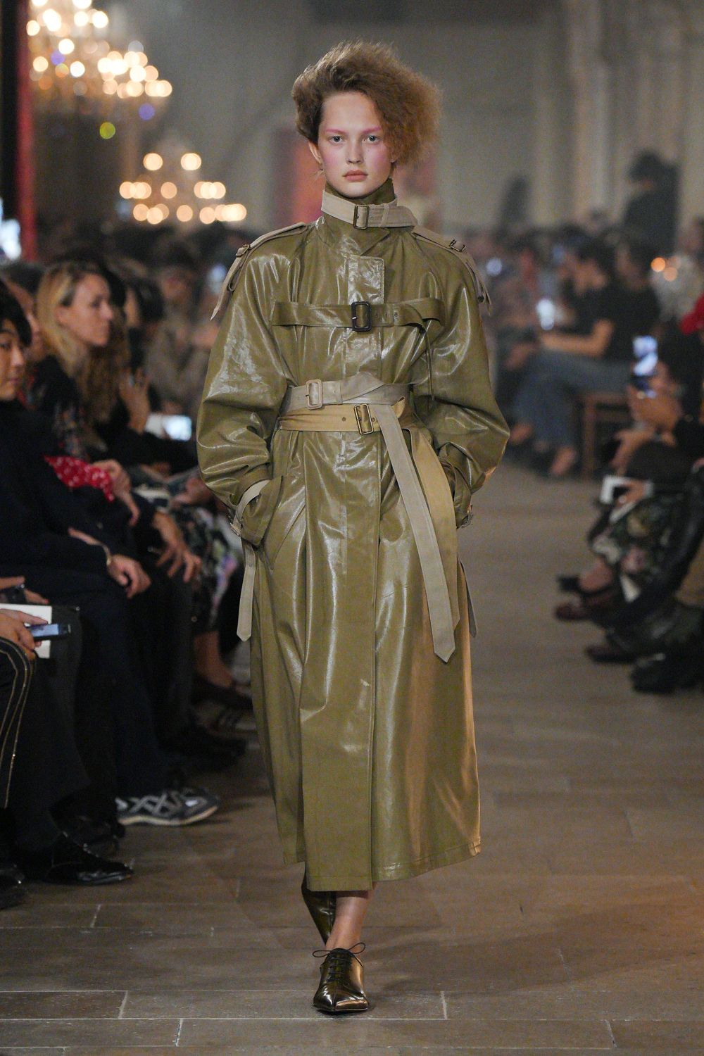 Olive glazed trench dress with throat latch, epaulettes, and doubled belts. Pointed olive lace-ups, one pale foot left bare. Whatever you wear crossing a drafty museum corridor after the rain.