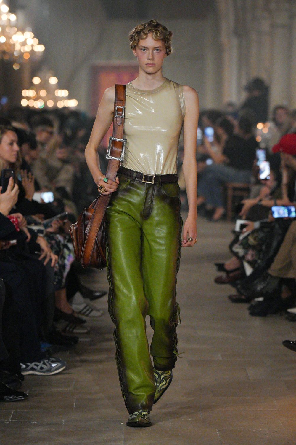 Creamy latex tank, stretched glossy across the chest. Oversized brown leather satchel on a wide buckled strap. Moss leather trousers with whipstitched seams snaking down the legs. Pointed boots in green scroll jacquard. Chunky rings, one set with a turquoise oval. Crossing a draughty hall after missing the hunt breakfast.