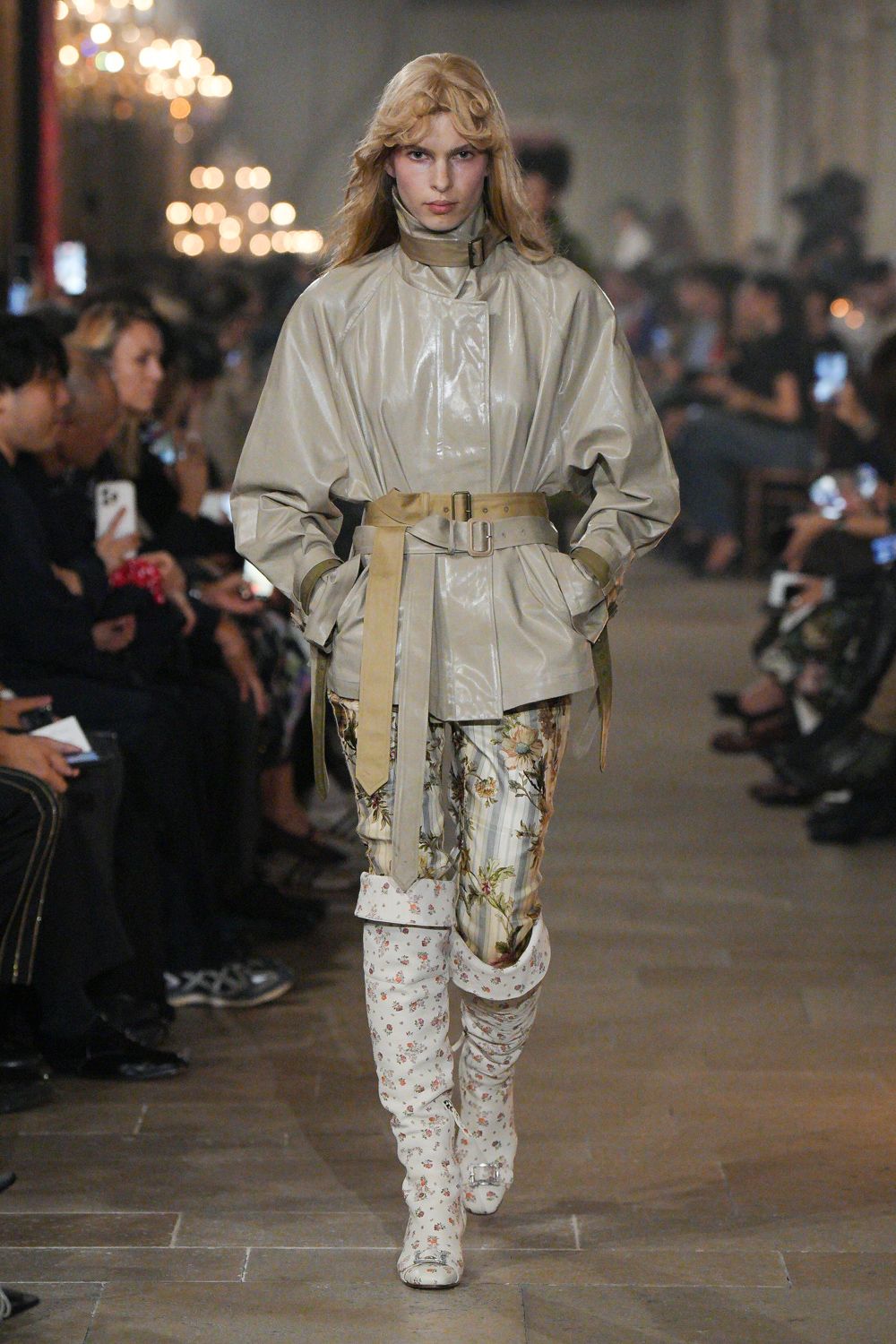 Pearl-grey coated jacket with a high buckled throat latch. Wide sand belt, doubled and hanging past the hip. Pale striped trousers overgrown with trailing cabbage-rose print. Cream floral boots ruched at the shin, square buckles at the toe. Whatever you wear home after sleeping through someone else's country-house weekend.