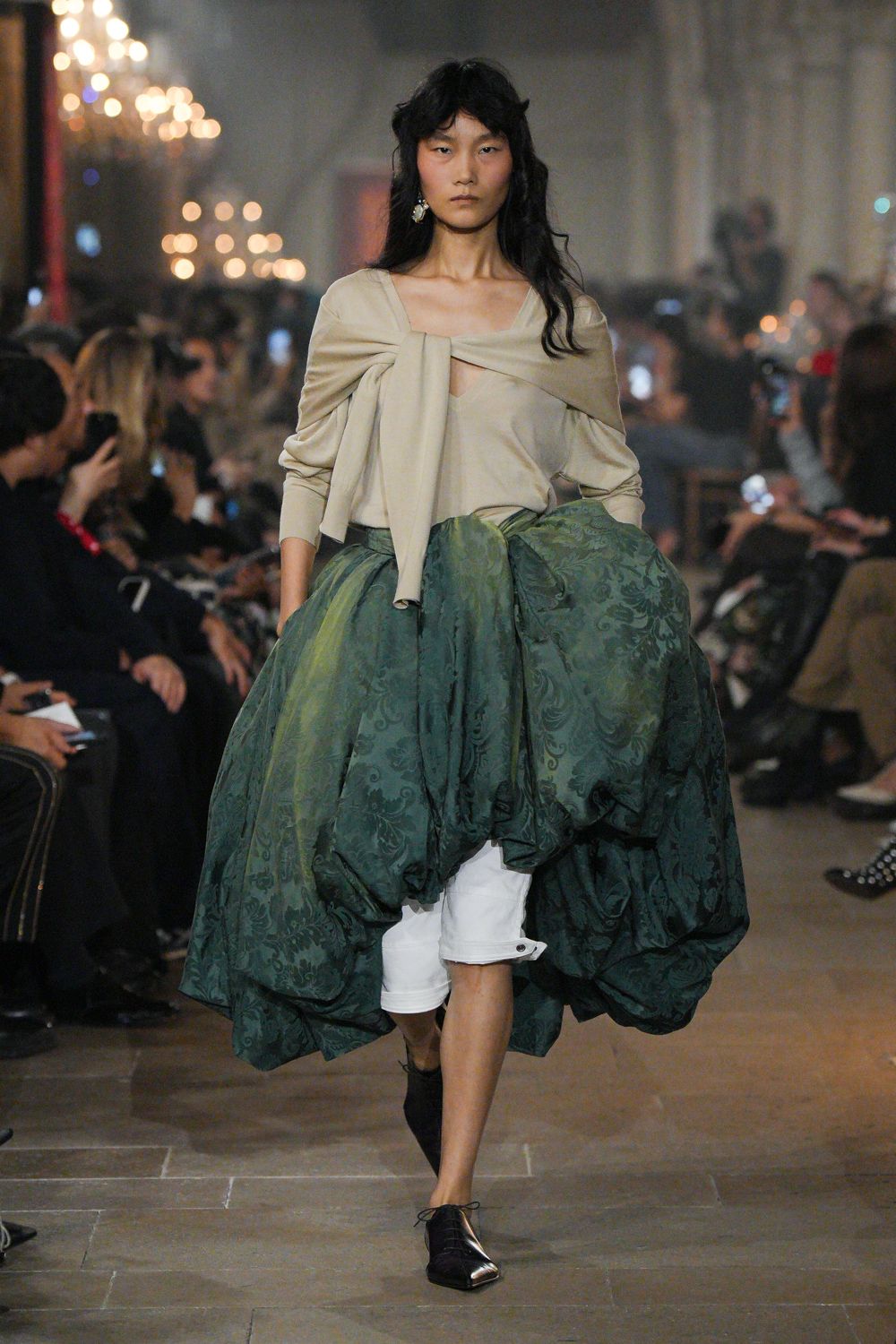 Oat draped top with a scarf panel knotted across the collarbones. Bottle-green jacquard overskirt, ballooned into uneven pannier-like volume. White knee-length shorts peeking cleanly beneath the lifted hem. Black square-toe lace-up flats. Crossing a drafty estate hallway after dinner, carrying half the tablecloth with you.