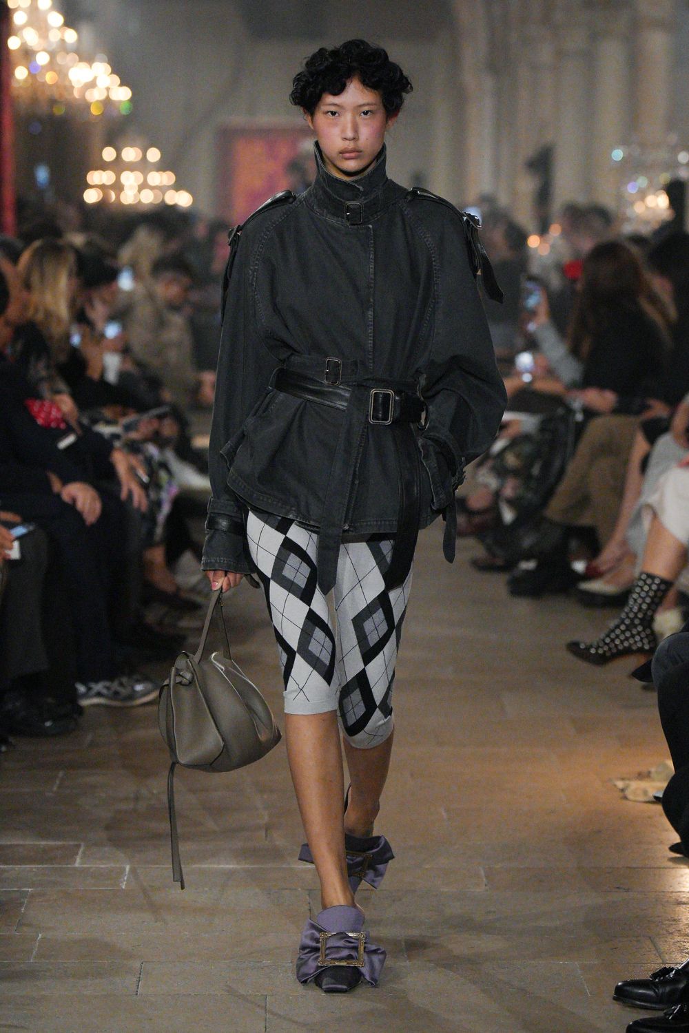 Charcoal washed jacket with a tall buckled collar and belted waist. White stretch cycling shorts crosshatched with black argyle lines. Taupe slouch bag with a long strap tail. Lavender satin mules gathered into bows under square buckles. Walking the hotel corridor after a wedding, carrying your shoes by midnight.
