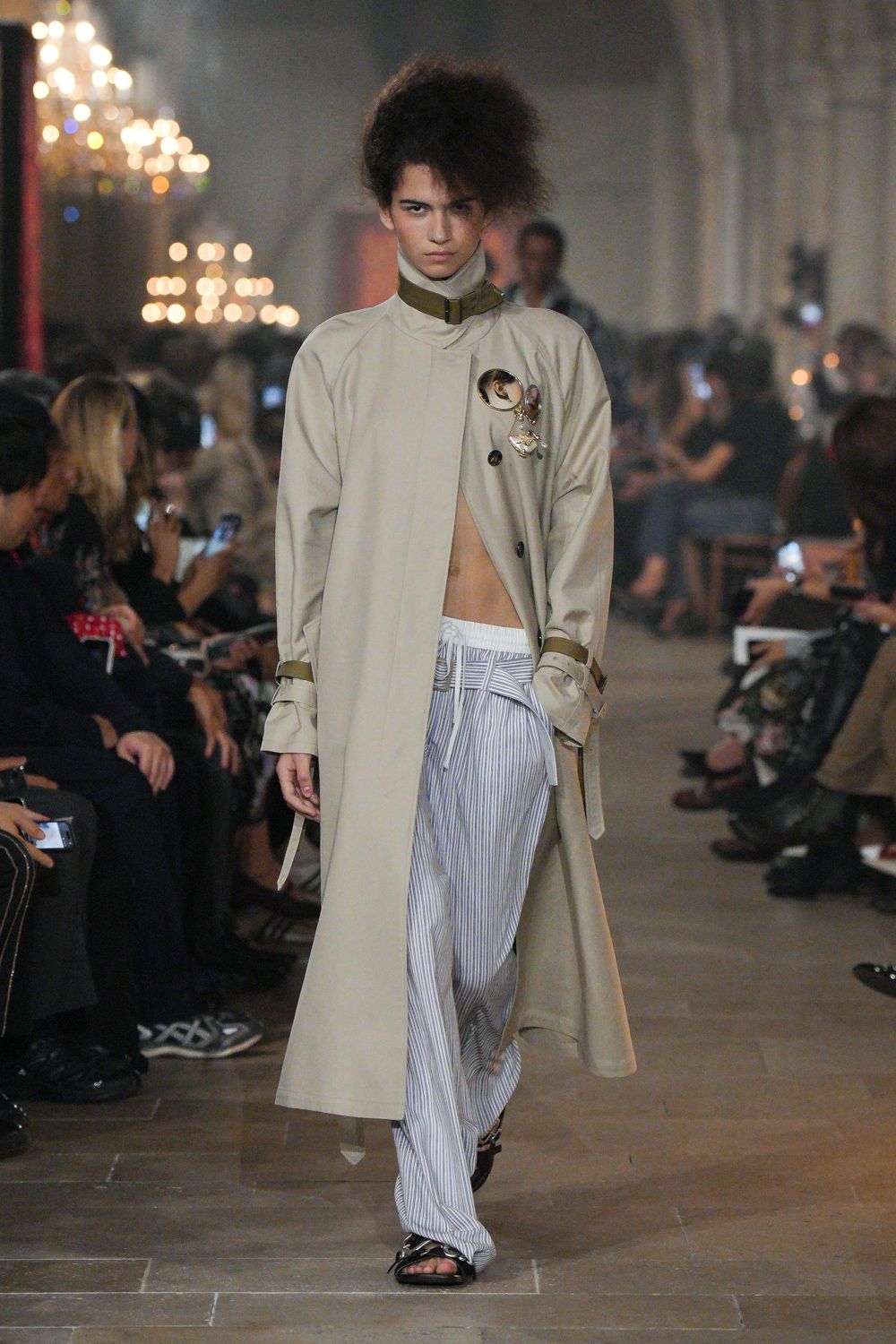 Pale stone trench coat with a buckled funnel neck and strap-cinched cuffs. Oversized cameo brooch pinned high on the chest. Blue-and-white striped drawstring trousers, waistband bunched above the hip. Black patent sandals with thin crossing straps. Whatever you throw on for dawn checkout in a grand hotel.