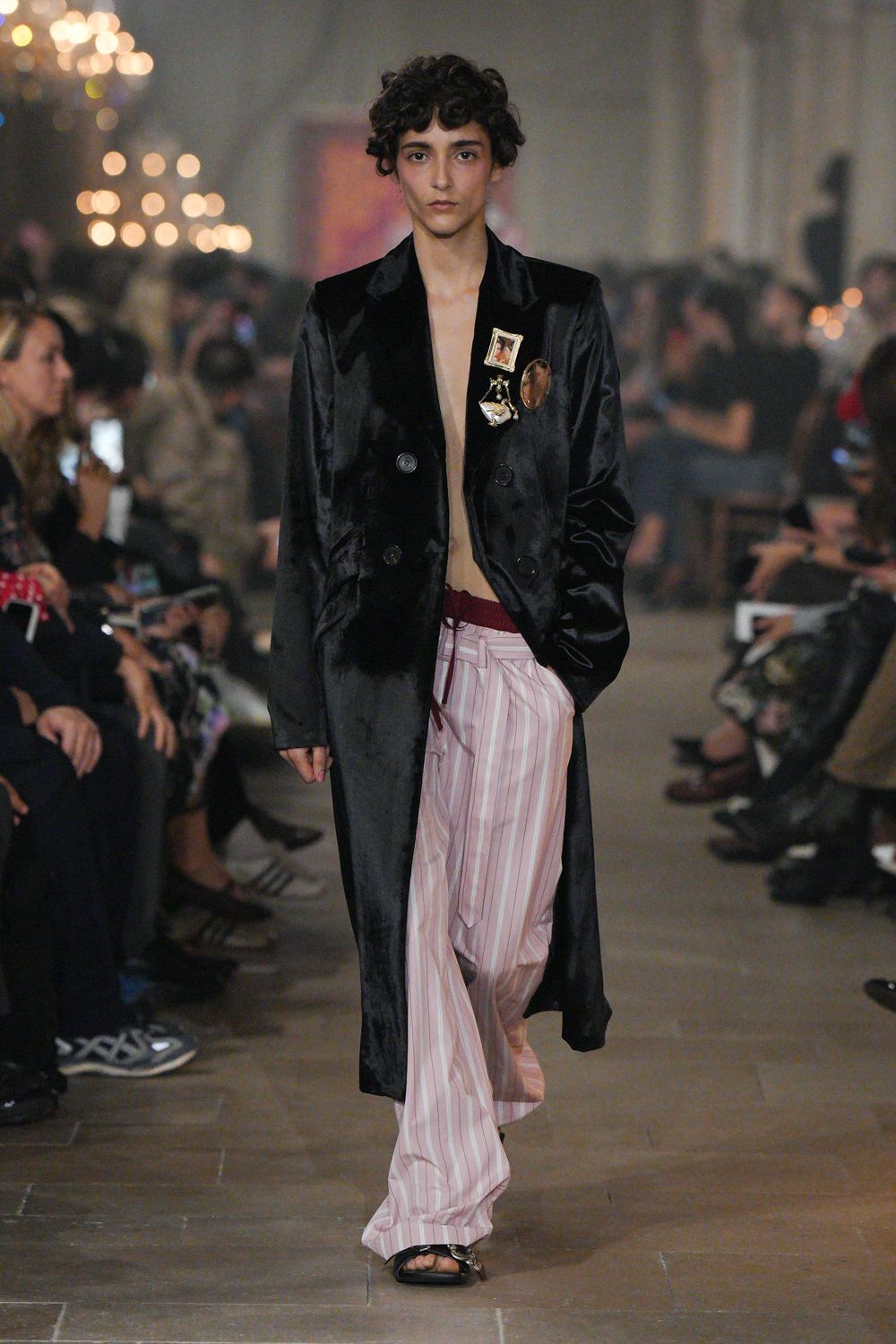 Black satin duster coat, double-breasted with souvenir pins on the lapel. Pale pink striped pyjama trousers, burgundy drawstring peeking above the waist. Flat black sandals with slim crisscross straps. Crossing a hotel corridor at dawn, still wearing last night's medal pins.