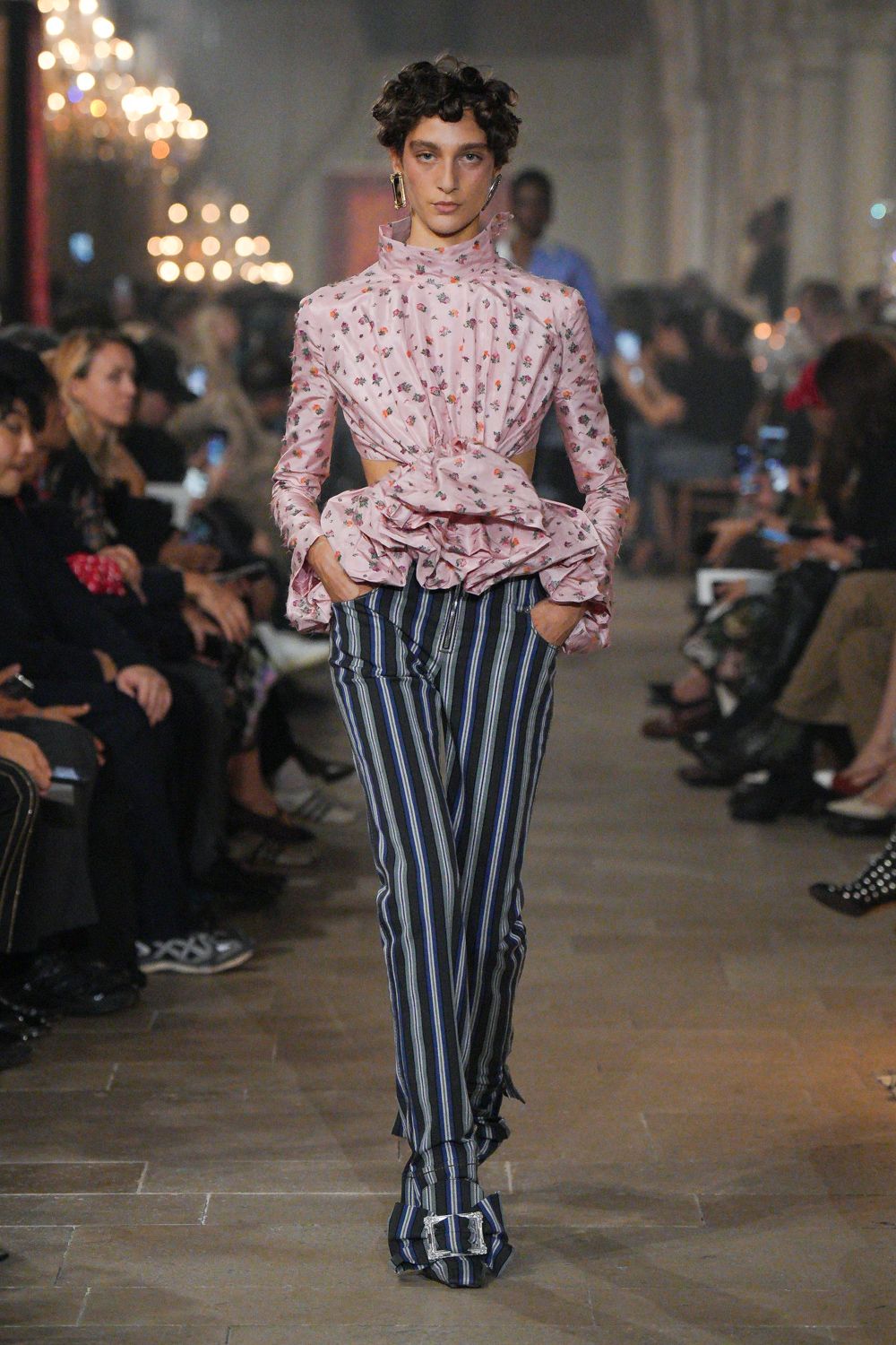 Blush floral blouse with a ruffled stand collar and huge tied peplum. Slim black trousers striped in white and blue, zipped at the front slit. Long rectangular metal drops. Matching striped shoes with squared silver buckles. Crossing a drafty palazzo ballroom after midnight champagne went flat.