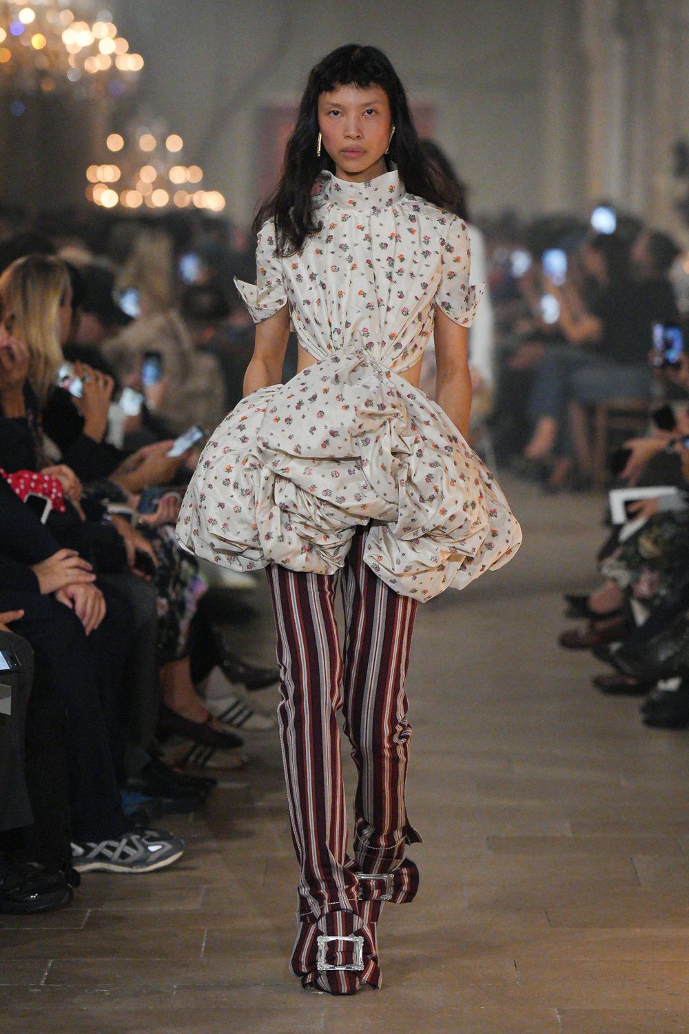 Ivory floral cotton dress, high neck and cutout waist knotted into a swollen bustle. Burgundy striped trousers, narrow through the leg with loose ankle ties. Matching striped boots with oversized square crystal buckles. Long gold bar earrings. Crossing a drafty manor corridor after pulling curtains off the guest bed.