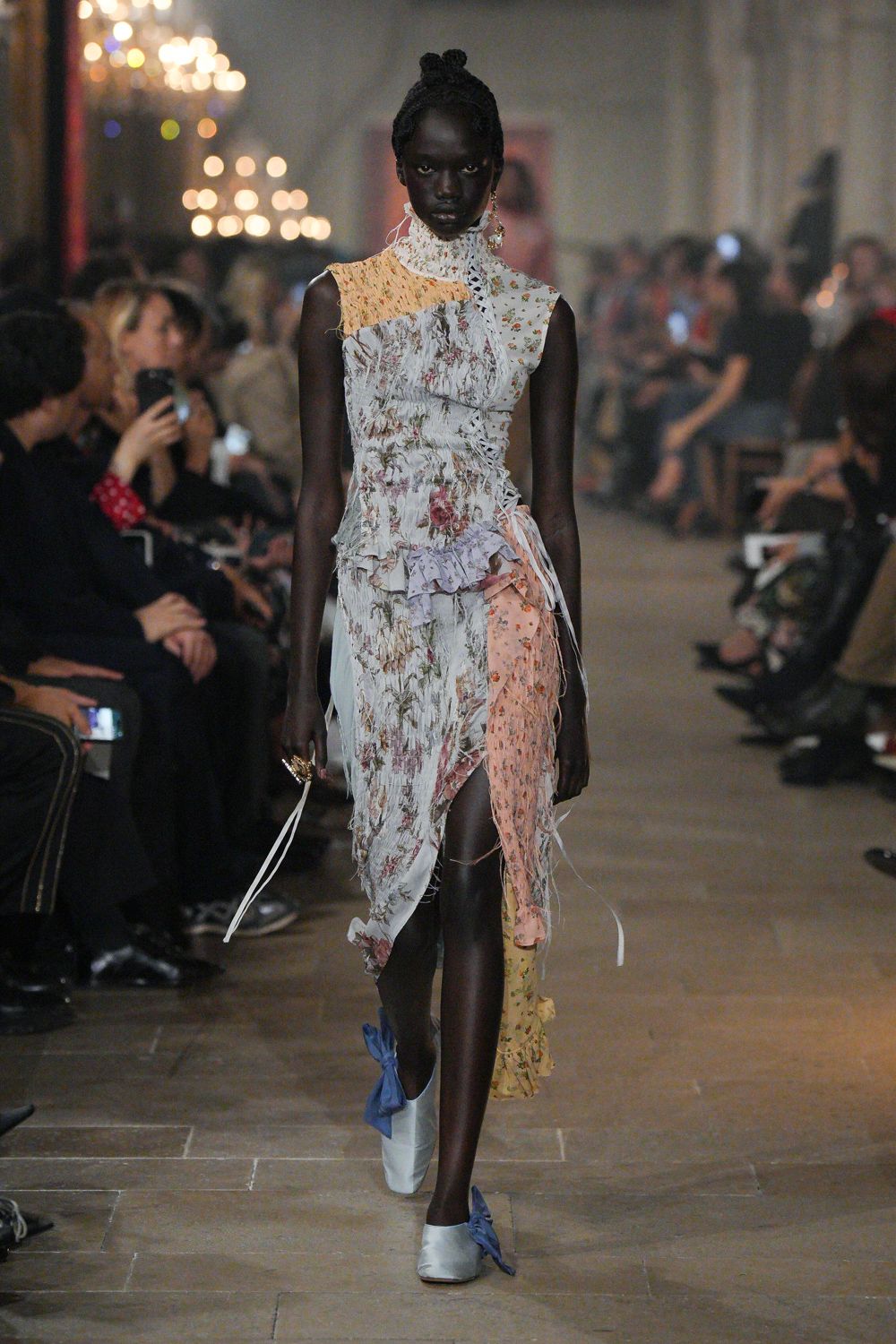 Patchworked floral dress in crinkled chiffon, ladder-laced through the torso. High white neck ruffle in broderie anglaise. Pale satin flats with oversized cornflower bows. Walking back from a wedding through wet grass at first light.