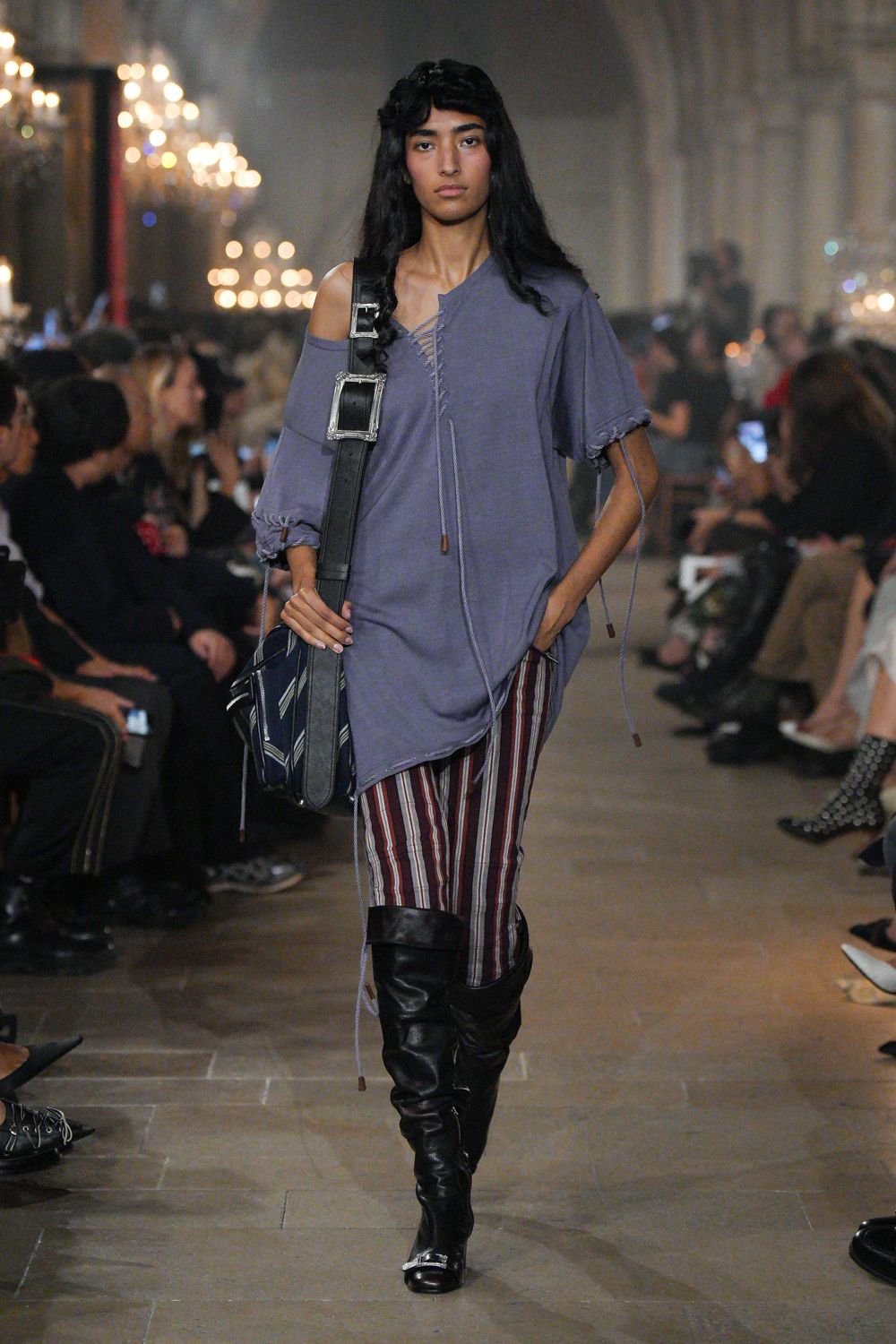 Washed violet jersey tunic, one shoulder dropped and seams left raw. Narrow trousers in burgundy and white vertical stripes. Black leather satchel on an oversized silver-buckled strap. Slouchy black knee boots with a metal-capped toe. Walking back from a village hall concert with the program still folded in your bag.