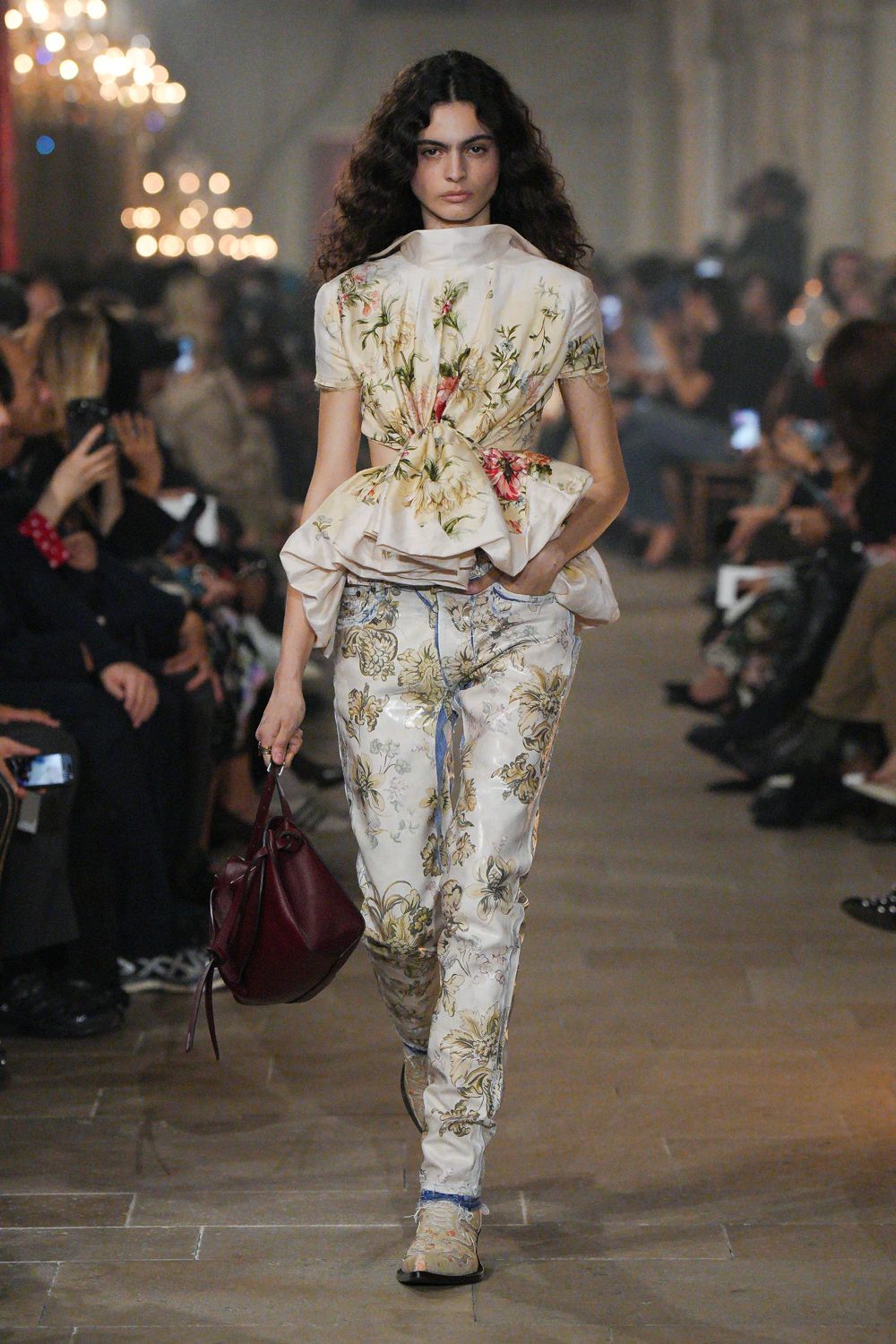 Ivory floral satin top with a high funnel neck and twisted peplum. Matching brocade trousers, blue seam piping and frayed hems breaking the shine. Pale floral ankle boots with raw edges at the opening. Burgundy slouch bag, thin strap dangling loose. Crossing a drafty manor corridor before dinner is announced.