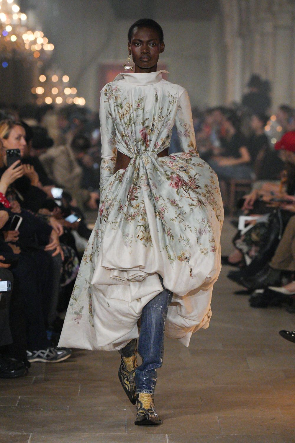 Ivory floral satin dress, funnel neck and vast side knot exposing the waist. Blue jeans with a silvered seam glint down one leg. Pointed python boots traced with dark curling overlays. Single dangling pearl cluster. Crossing a draughty manor corridor after midnight, carrying half the curtains with you.