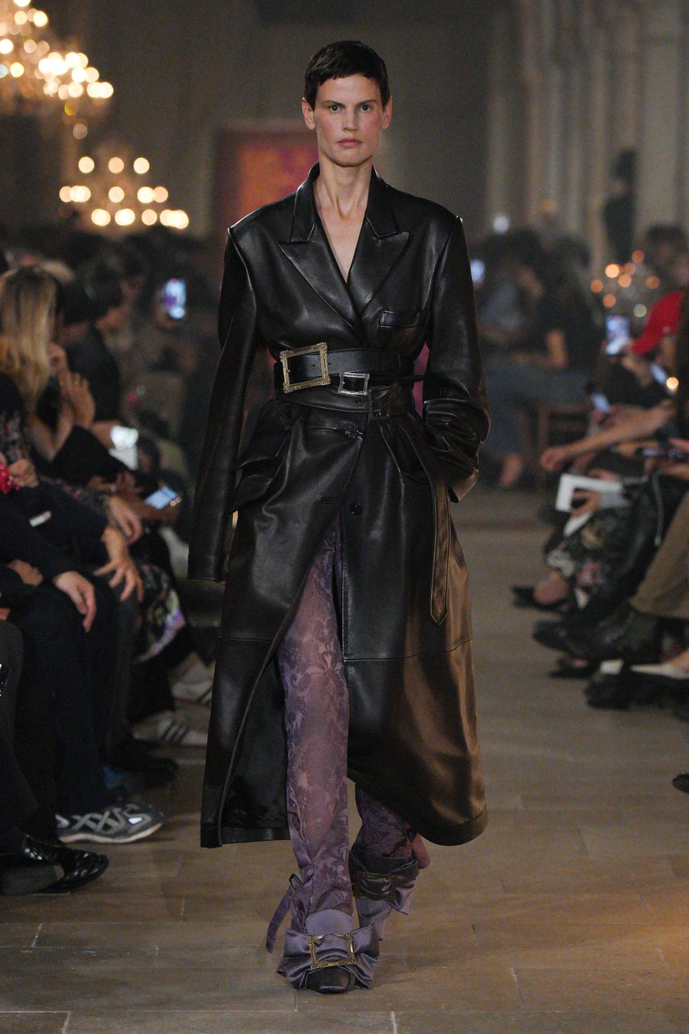 Black leather coatdress with broad lapels and a low wrapped front. Stacked black leather belts, one buckle oversized and gilt-framed. Mauve jacquard trousers pooling into soft folds at the ankle. Square-toe pumps strapped with matching mauve fabric. Whatever you wear to cross a candlelit hall after midnight.