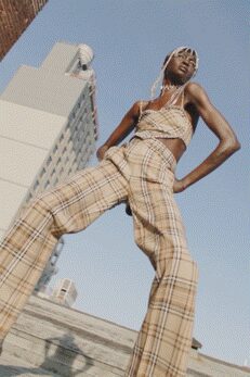 Pearled net headscarf draped close over the hairline. Crocheted halter top in open ivory squares, tied under the bust. High-waisted beige plaid flares breaking wide from the knee. On sun-blasted steps outside a hotel where the pavement throws the heat back.