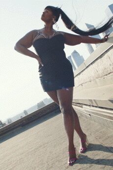 Black ruched bodycon mini with a sweetheart neckline edged in white piping. Short silver chain at the collarbone. Small hoop earrings. Hot-pink heeled sandals with narrow glossy straps. On a rooftop parking deck, wind pulling the ponytail sideways.
