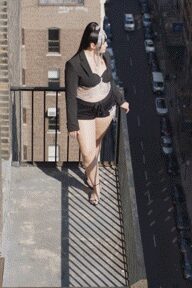 Black ribbed cardigan slipping off one shoulder, fastened with a single button. Black balconette bra with scalloped edges and a tiny center bow. High-cut black briefs ruched across the front. Sheer nude tights catching the sun on the shin. Black ankle-strap heels. On a fire escape at noon, traffic grinding far below.