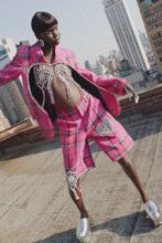 Hot-pink blazer hanging open over a pearl-swirled bare chest. Pink plaid knee shorts with a white panel wrapping one thigh. White slip-ons, clean and blunt at the toe. Single drop earring. On a hotel roof at noon, wind catching the open jacket first.