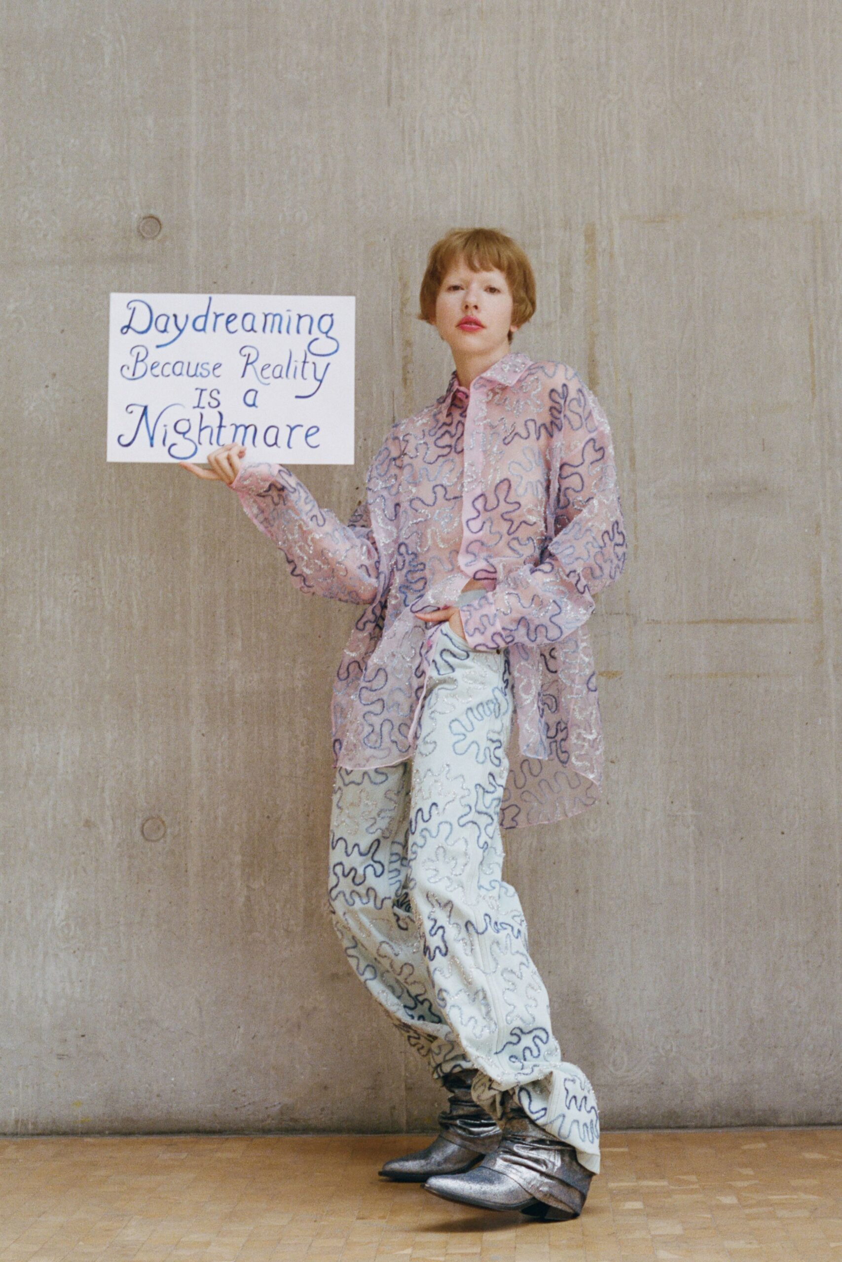 Oversized blush shirt traced with navy squiggle embroidery and scattered sequins. Cream wide-leg trousers with the same looping embroidery, pooling at the ankles. Silver pointed boots with a crumpled metallic shaft. Holding a homemade placard in a concrete lobby before the meeting starts.