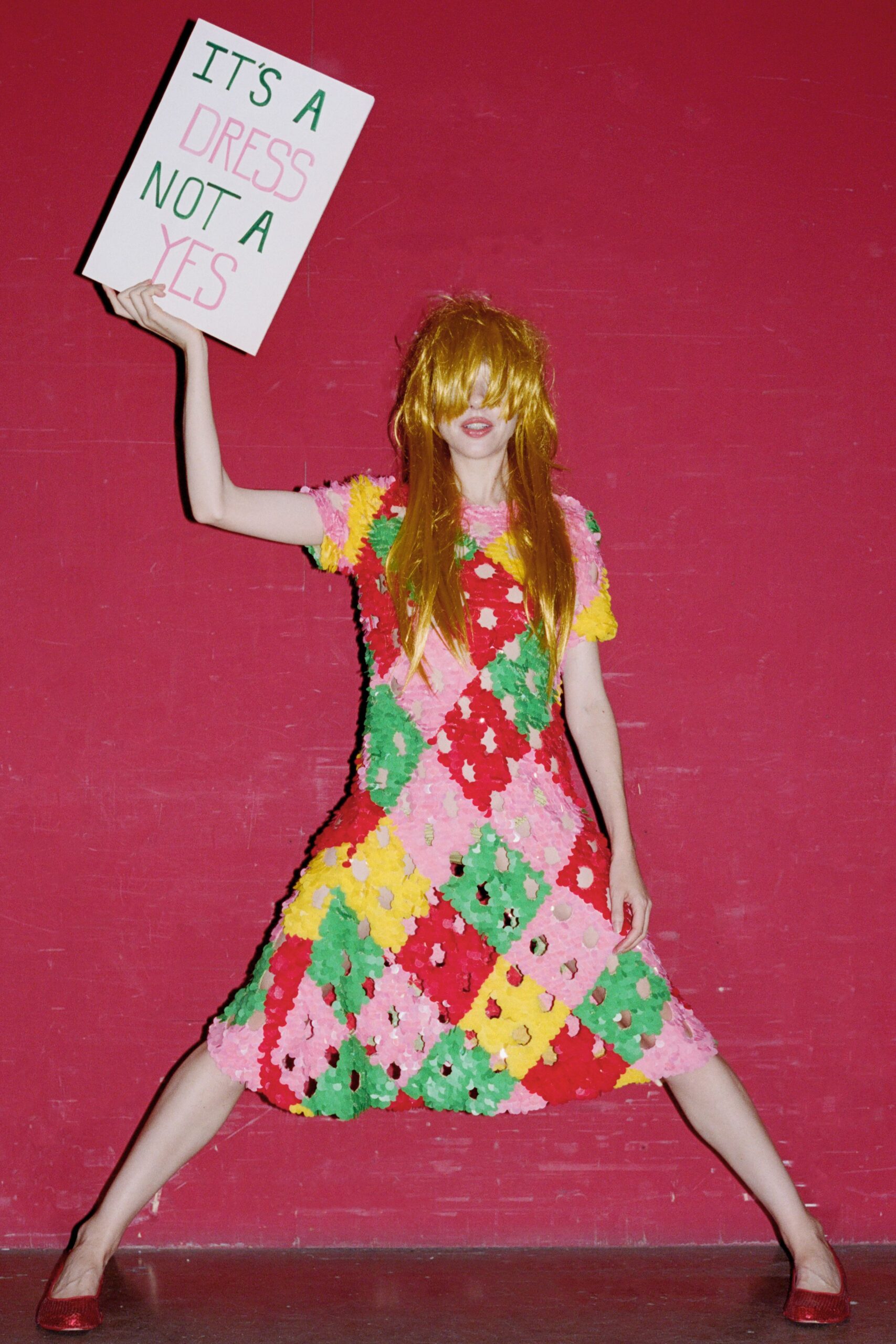 Long metallic-gold wig falling over the eyes in uneven strands. Fit-and-flare dress built from petal-like paillettes in pink, red, yellow and green. Red glitter flats with a rounded toe. Holding a protest placard in a community hall after midnight.