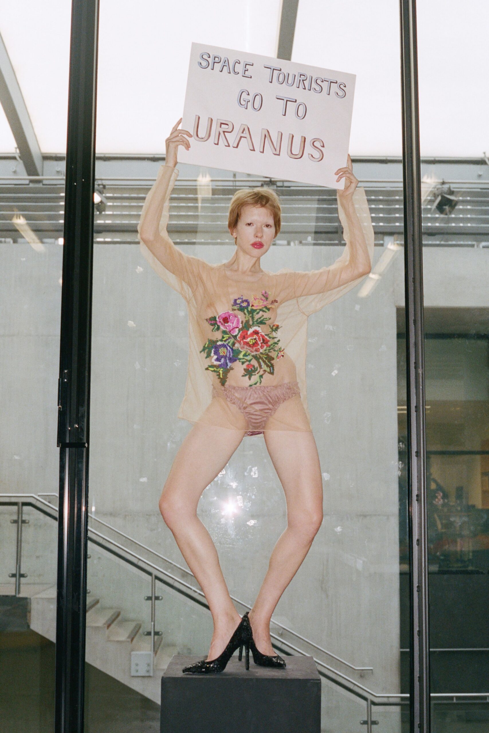 Nude mesh top with a dense cross-stitched bouquet across the torso. Mauve satin briefs, ruched and high-cut at the hip. Black sequined pumps with narrow stiletto heels. Standing on a plinth at a student protest inside the atrium.