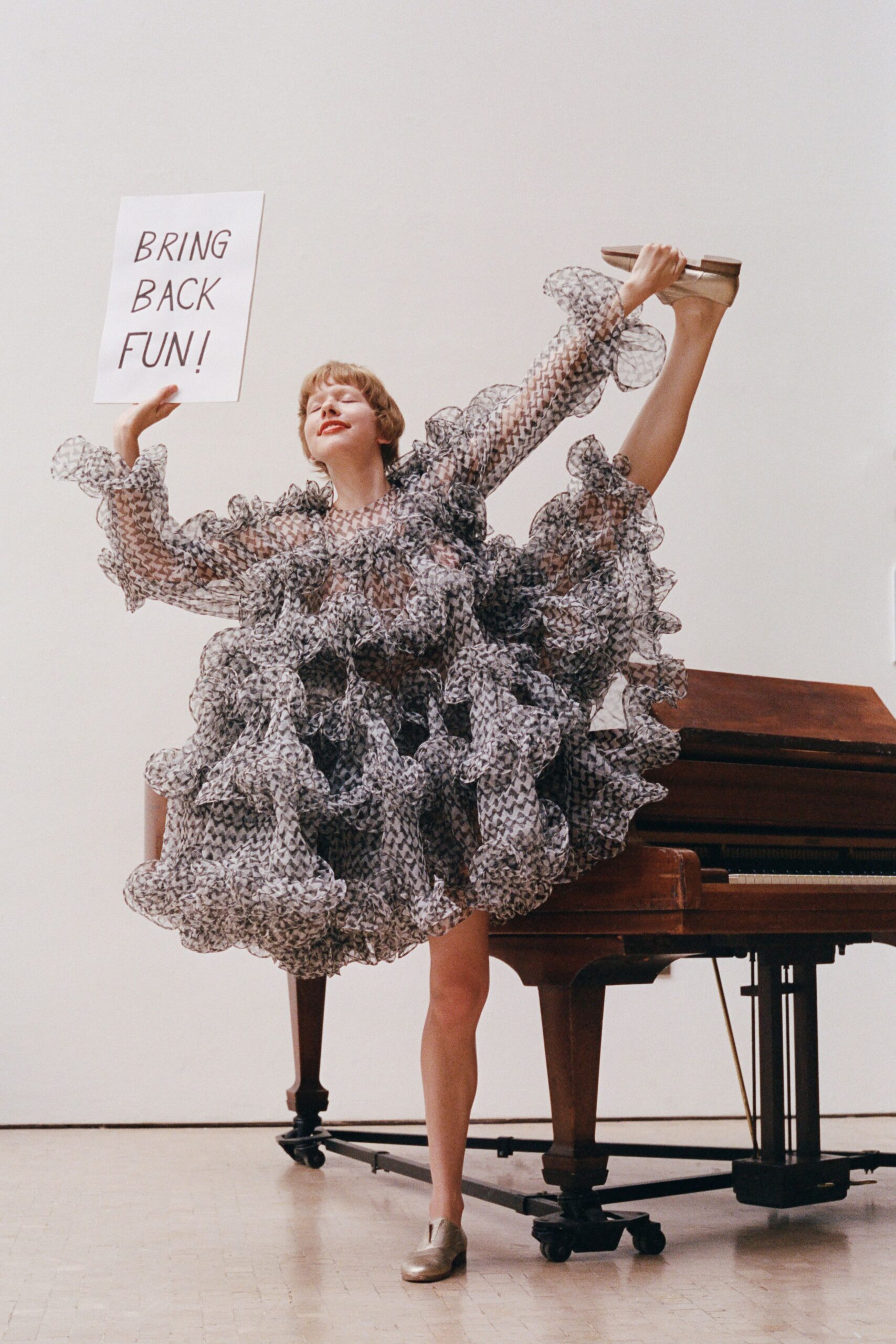 Sheer black-and-white chiffon dress, ballooned with dense wired ruffles and zigzag print. Champagne leather flats with squared toes and a low stacked heel. Climbing onto the rehearsal piano after somebody taped demands to the wall.