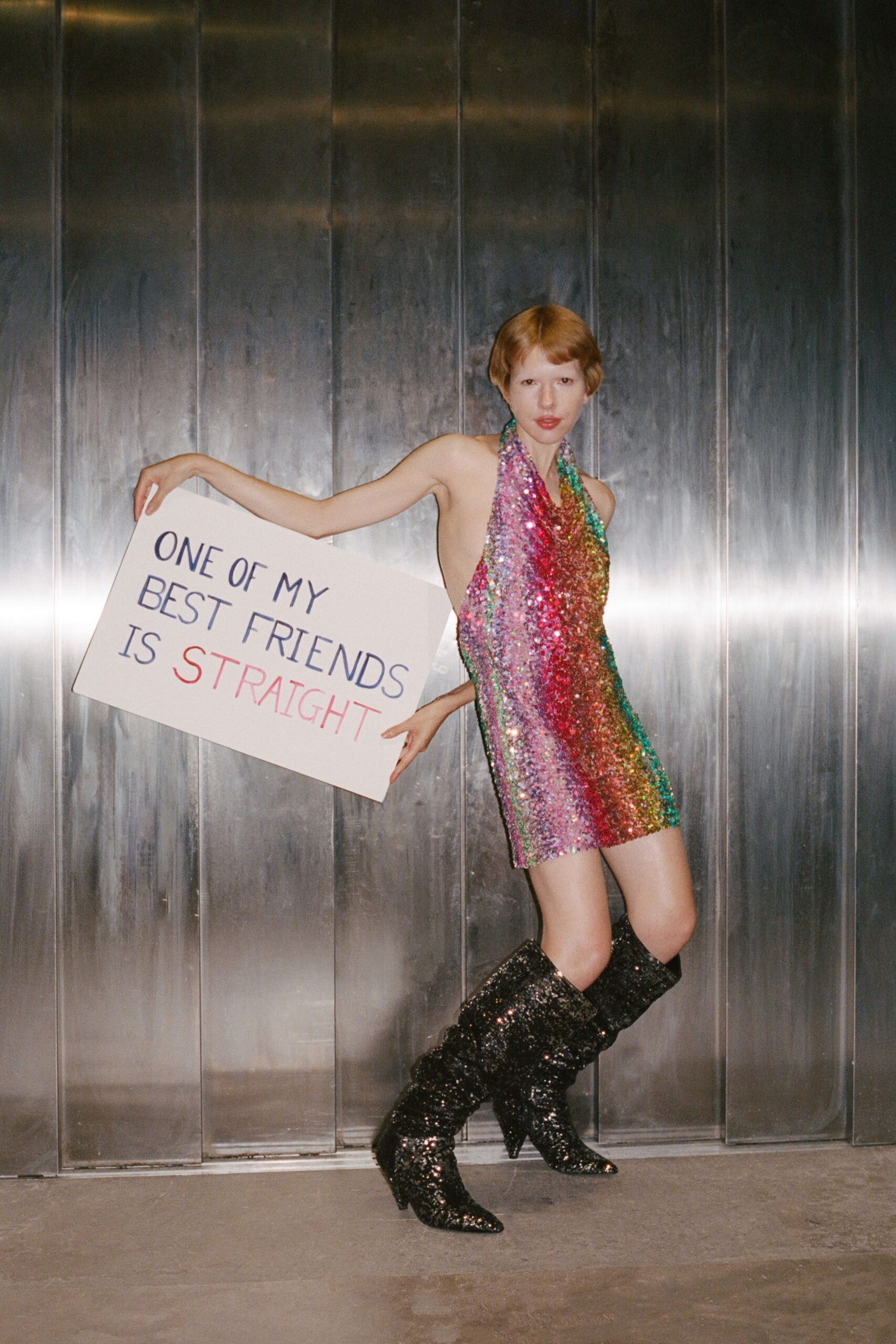 Rainbow paillette halter mini, side cut to bare nearly the whole torso. Black sequined boots slouched at the shin over narrow stiletto heels. Holding a protest placard outside the club while the smokers drift back in.
