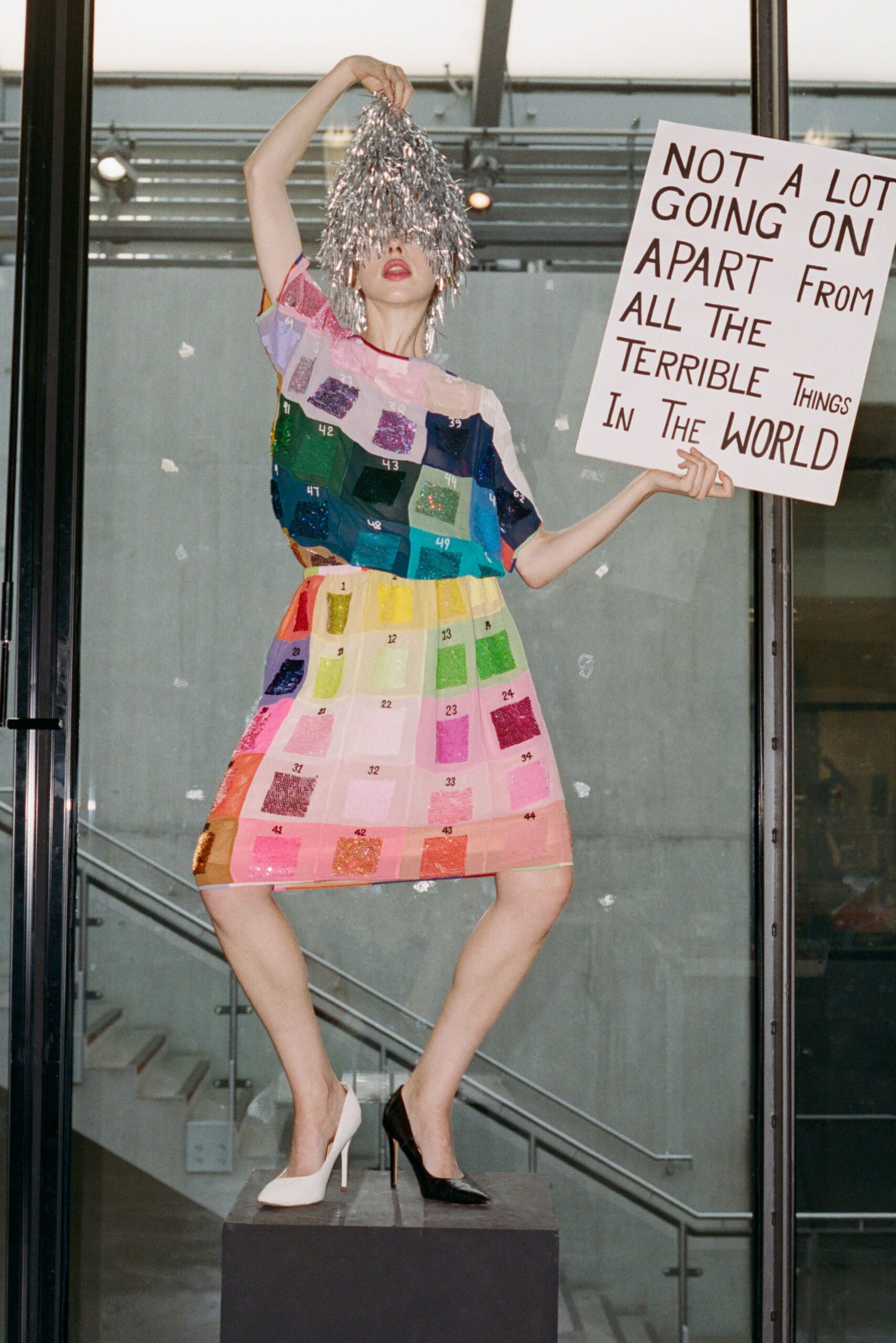 Silver foil tinsel hood falling over the eyes like a mop. Sheer short-sleeved shift printed as a numbered color chart, glitter swatches stitched in grids. Mismatched leather pumps, one white and one black. Holding a protest sign at the museum atrium after the opening speech soured.