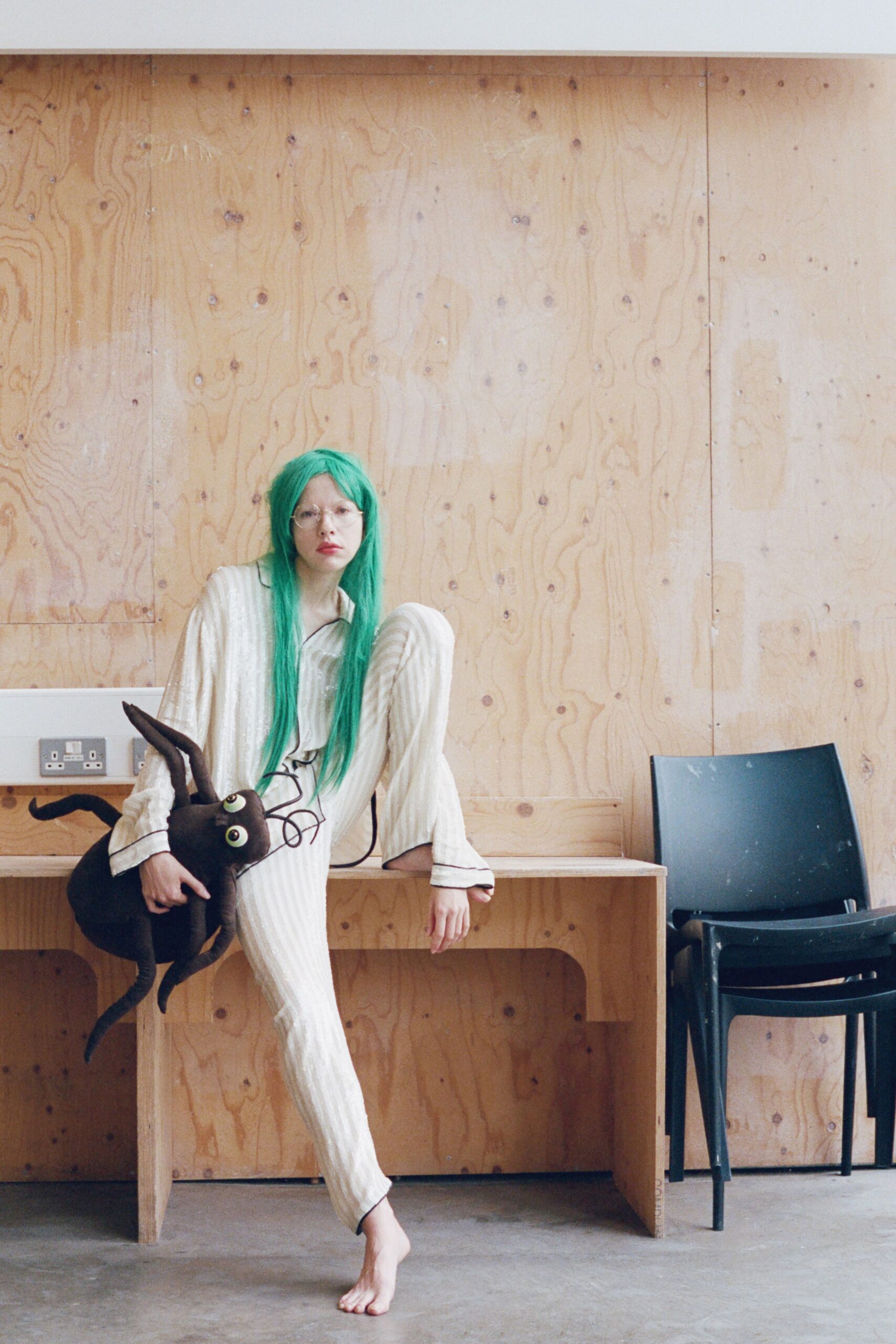 Cream crinkled pajama shirt with black piping tracing the collar. Matching pleated trousers, one leg pooling softly over the bare foot. Round wire glasses. Stuffed black cat bag with bulging eyes and loose cord whiskers. Waiting in a rented rehearsal hall after someone misplaced the house keys.