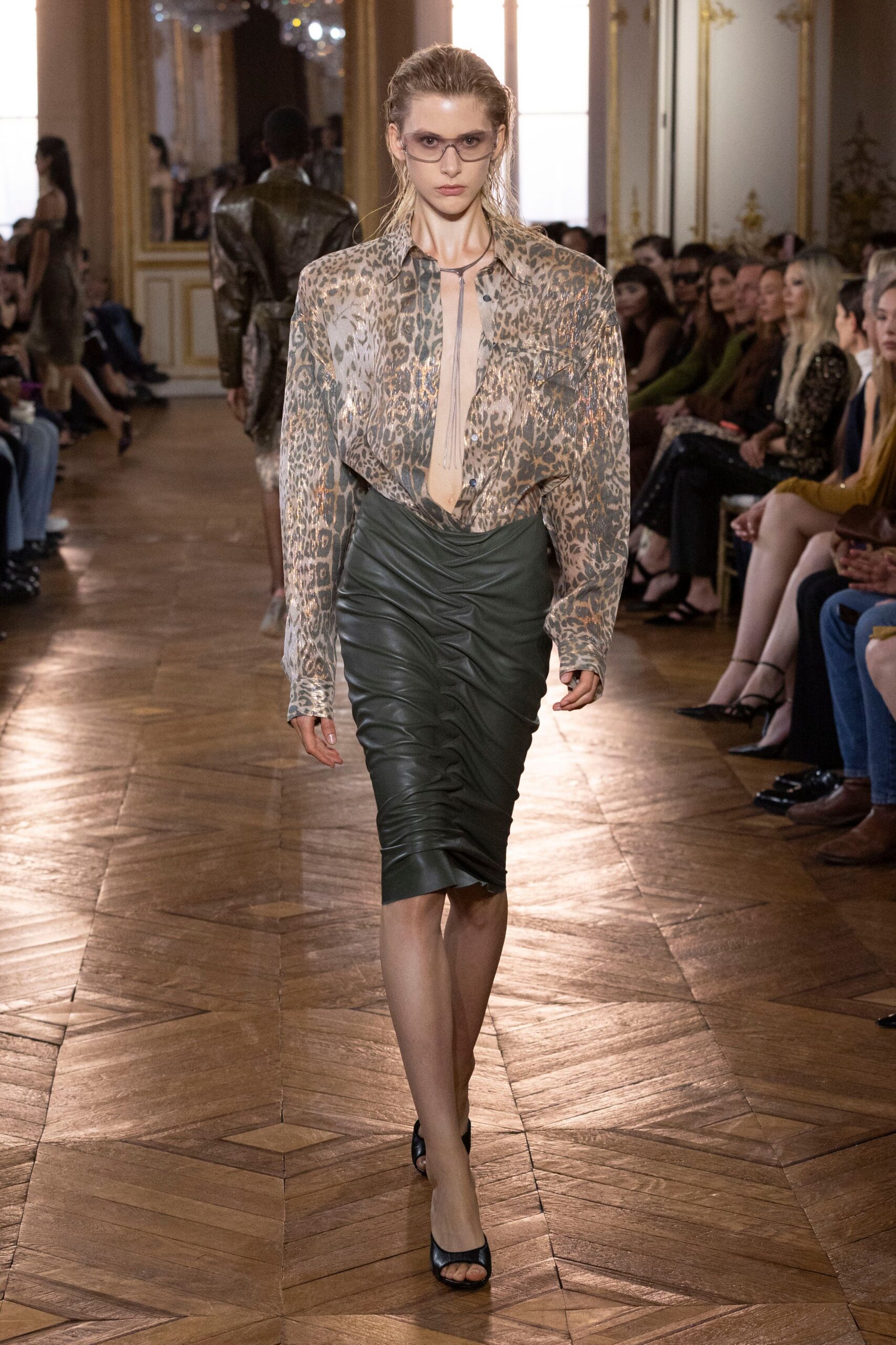 Smoked wraparound glasses with a narrow silver bridge. Sheer leopard chiffon shirt washed with bronze shine, unbuttoned to the navel. Long silver lariat dropping straight into the plunge. Slate leather pencil skirt, densely ruched down the front. Black peep-toe mules. Hotel bar at noon, still wearing last night's hair.