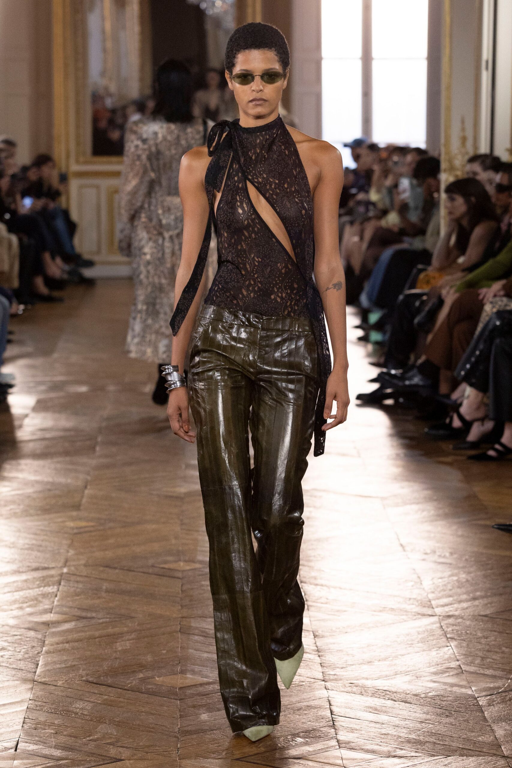 Narrow olive-tinted lenses in a slim metal frame. Black lace halter, one shoulder tied off with a long ribbon. Olive crocodile-embossed leather trousers with a low curved waistband. Stacked silver cuffs. Pale sage pointed pumps. Whatever you wear to miss the last train and keep going.