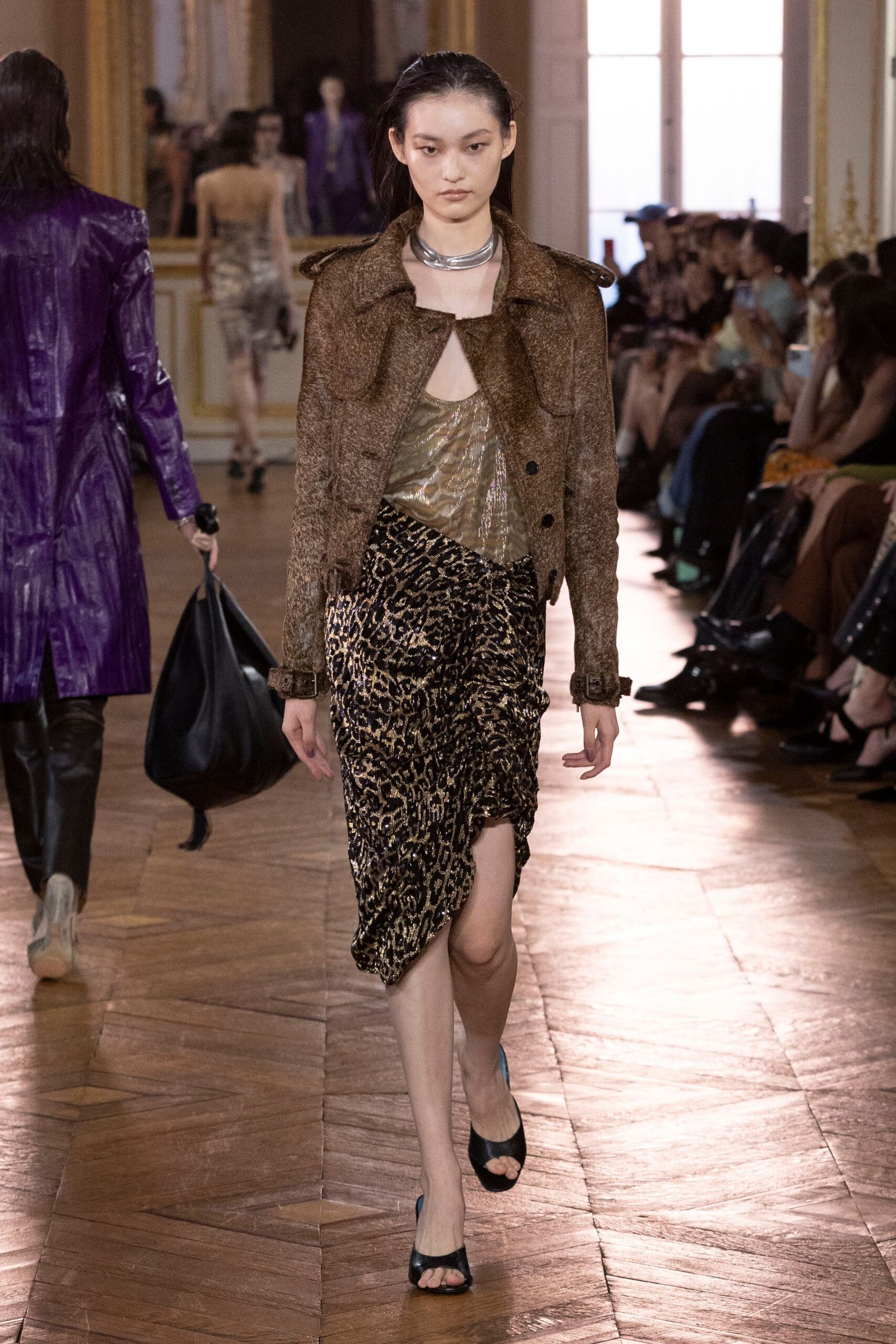 Stacked silver torque hugging the collarbones. Brown tweed cropped jacket with epaulettes and buckled cuffs. Olive metallic camisole, zebra shimmer pooling under the bust. Black and gold leopard wrap skirt, ruched into a high slit. Black peep-toe mules. Crossing a parquet ballroom at the hour the champagne turns flat.