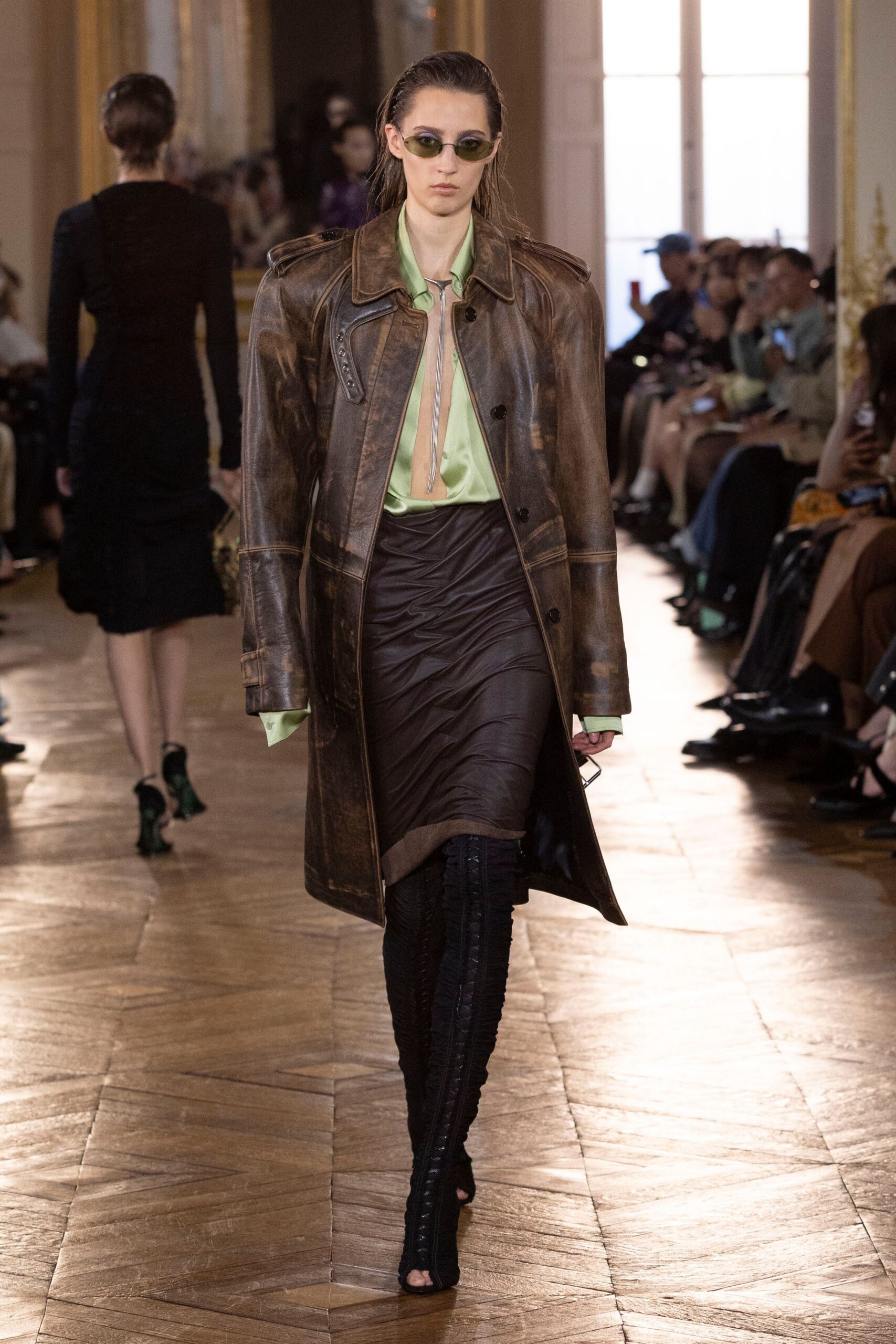 Narrow olive lenses in a thin metal frame. Weathered brown leather coat with epaulettes and bleached wear marks. Pale pistachio satin blouse hanging open to a deep slit. Dark ruched skirt, twisted across the hips. Black lace-up sock boots with open toes. Walking into city hall after sleeping in yesterday's clothes.