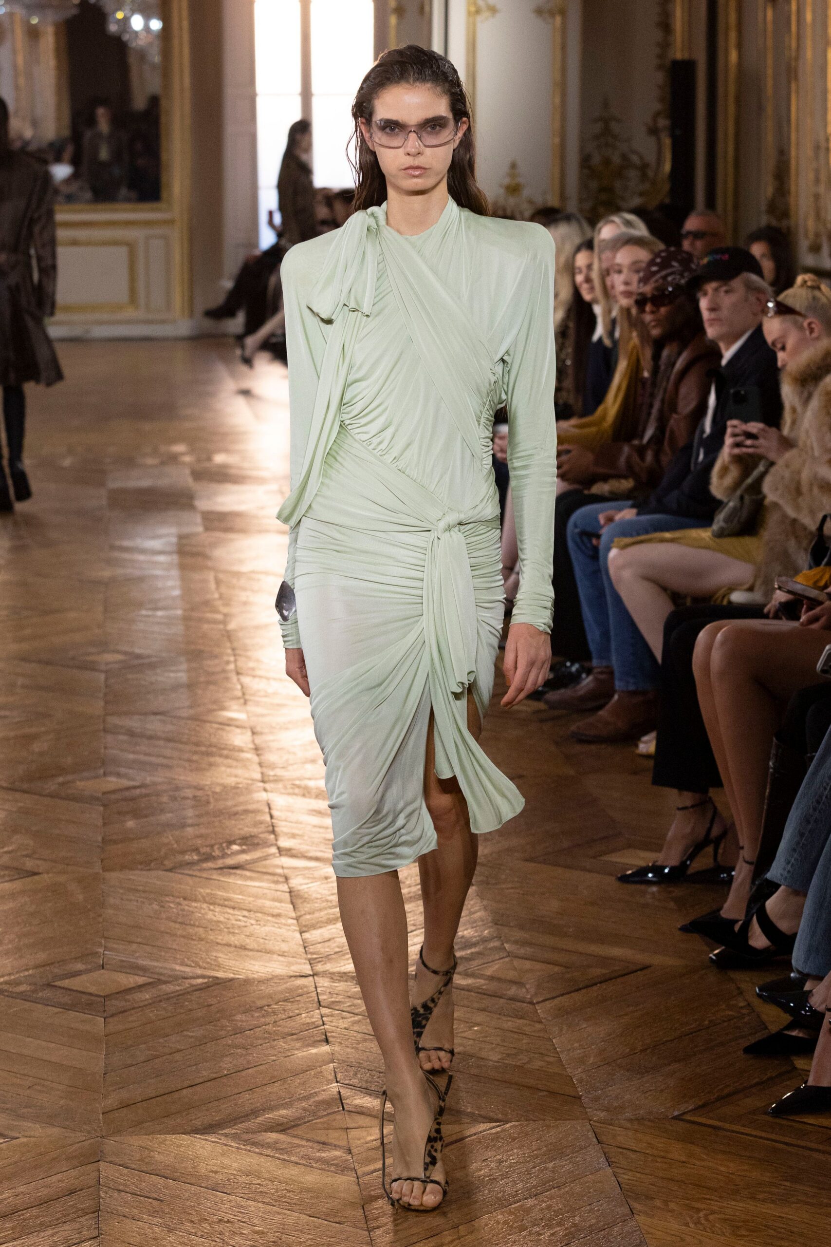 Smoke-tinted shield sunglasses with a narrow wraparound line. Pale mint jersey dress, ruched and cross-draped into a knotted front slit. Leopard calf-hair stiletto sandals with barely there straps. Crossing a hotel lobby before breakfast, still carrying last night's makeup.