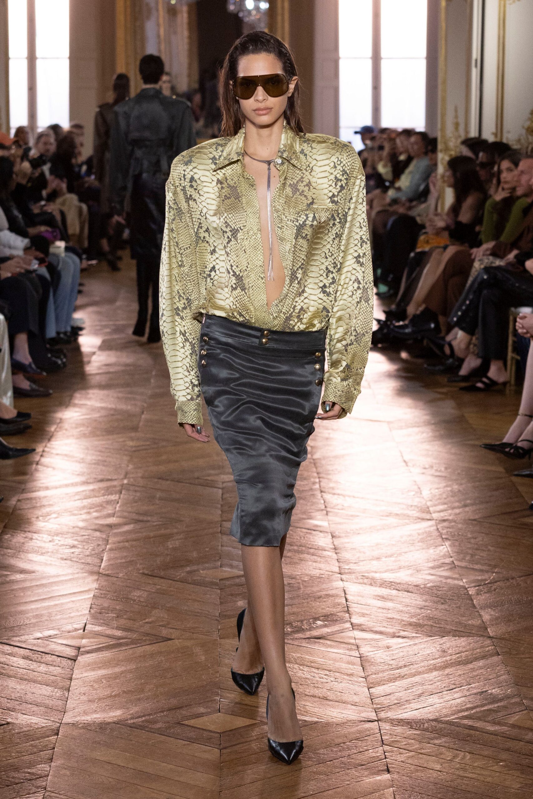 Oversized amber visor sunglasses. Olive python-print satin blouse, plunged open to the waist. Long silver lariat ending in a narrow tassel. Charcoal satin pencil skirt, double rows of brass side buttons. Black pointed leather pumps. Hotel ballroom at noon, parquet glowing after the rain.