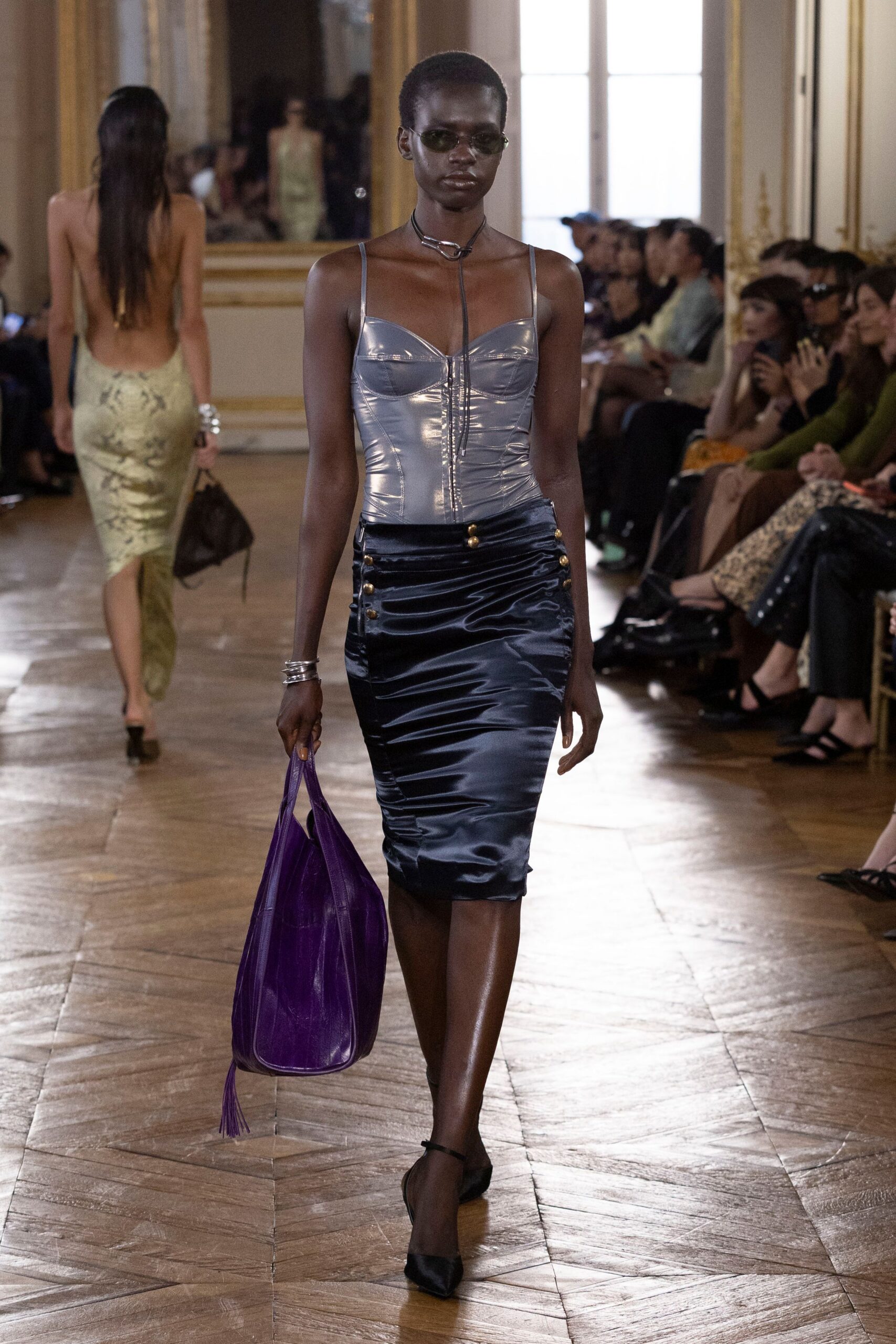 Narrow olive-tinted oval lenses. Black cord lariat with a chunky silver ring. Steel-blue coated corset, ruched through the zip front. Ink satin pencil skirt, brass buttons marching from the hips. Oversized violet leather hobo with a long tassel. Black satin slingbacks. Walking into a hotel bar before the rain has let up.