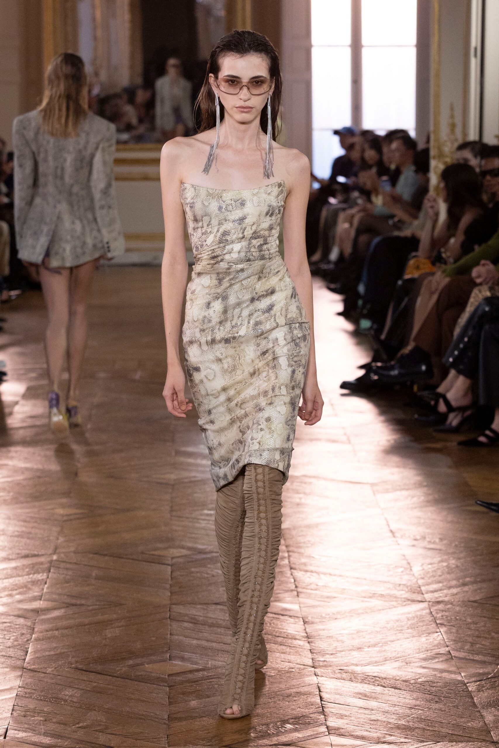 Light brown tinted aviators with a thin metal rim. Long silver fringe earrings brushing the collarbone. Strapless snakeskin-print dress, tightly ruched through the waist and hip. Taupe ruched thigh-high boots with a laced strip down the front. Crossing a hotel atrium before breakfast, still in last night's makeup.