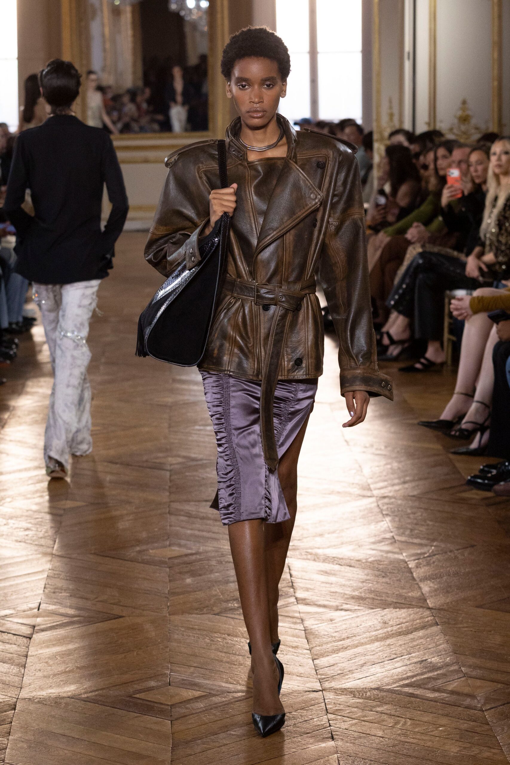 Oversized brown leather biker jacket, belted waist and sloped padded shoulders. Stacked gunmetal torque rings at the throat. Black suede hobo bag with a silver-edged panel. Lilac satin pencil skirt, ruched into a sharp thigh slit. Black pointed pumps. Whatever you wear to cross a hotel lobby before sunrise.