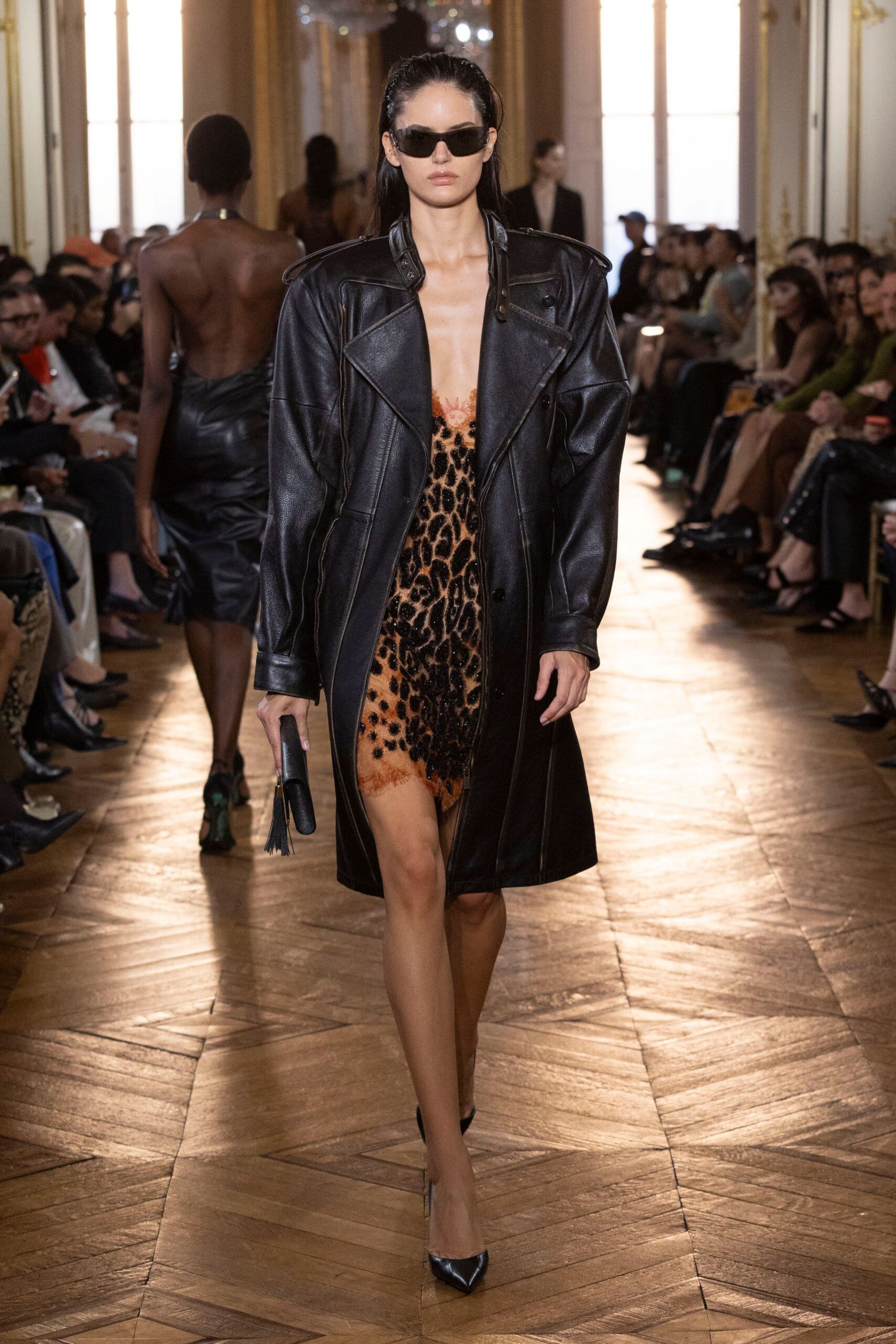 Narrow black wraparound sunglasses. Oversized black leather coat with dropped shoulders and epaulets curling upward. Nude leopard slip, beaded black spots and rust lace at the neckline. Black tube clutch with a tassel swing. Black pointed pumps. Hotel corridor after midnight, keycard bent in the bottom of the bag.