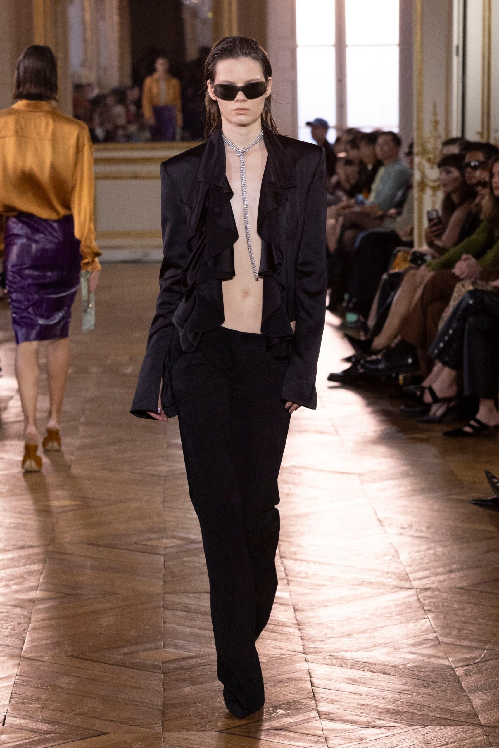 Narrow black wraparound sunglasses. Rhinestone lariat dropping straight down the bare sternum. Black jacket cut open to the waist, lapels falling in soft ruffles. Fluid black trousers with a long break over the shoe. Crossing a hotel ballroom floor before anyone has cleared the glasses.