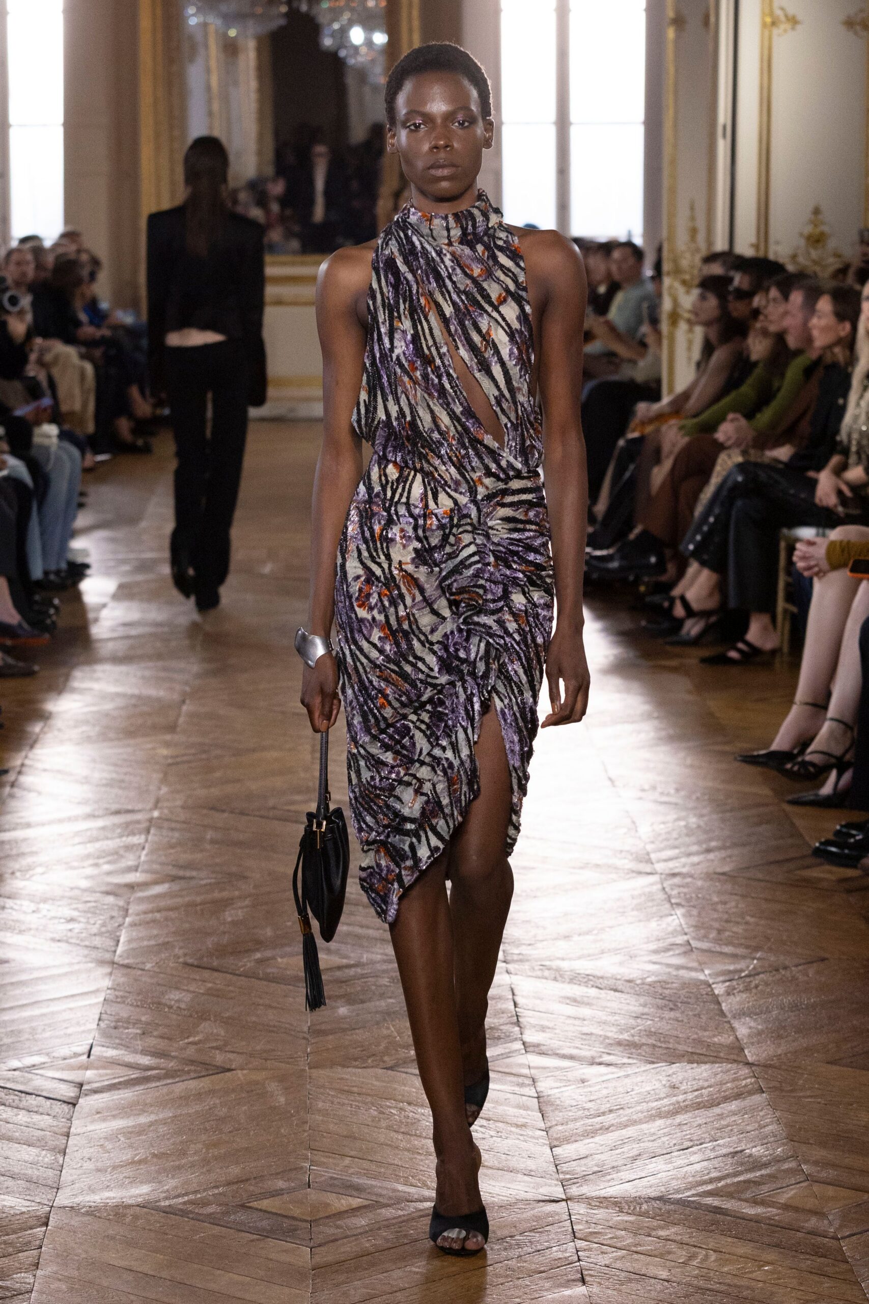 Ruched halter dress in painterly velvet devore, slit opening from bust to thigh. Wide silver cuff, bent like poured metal. Black tassel pouch with a thin wrist strap. Black peep-toe mules. Crossing a hotel ballroom after midnight with one earring missing.