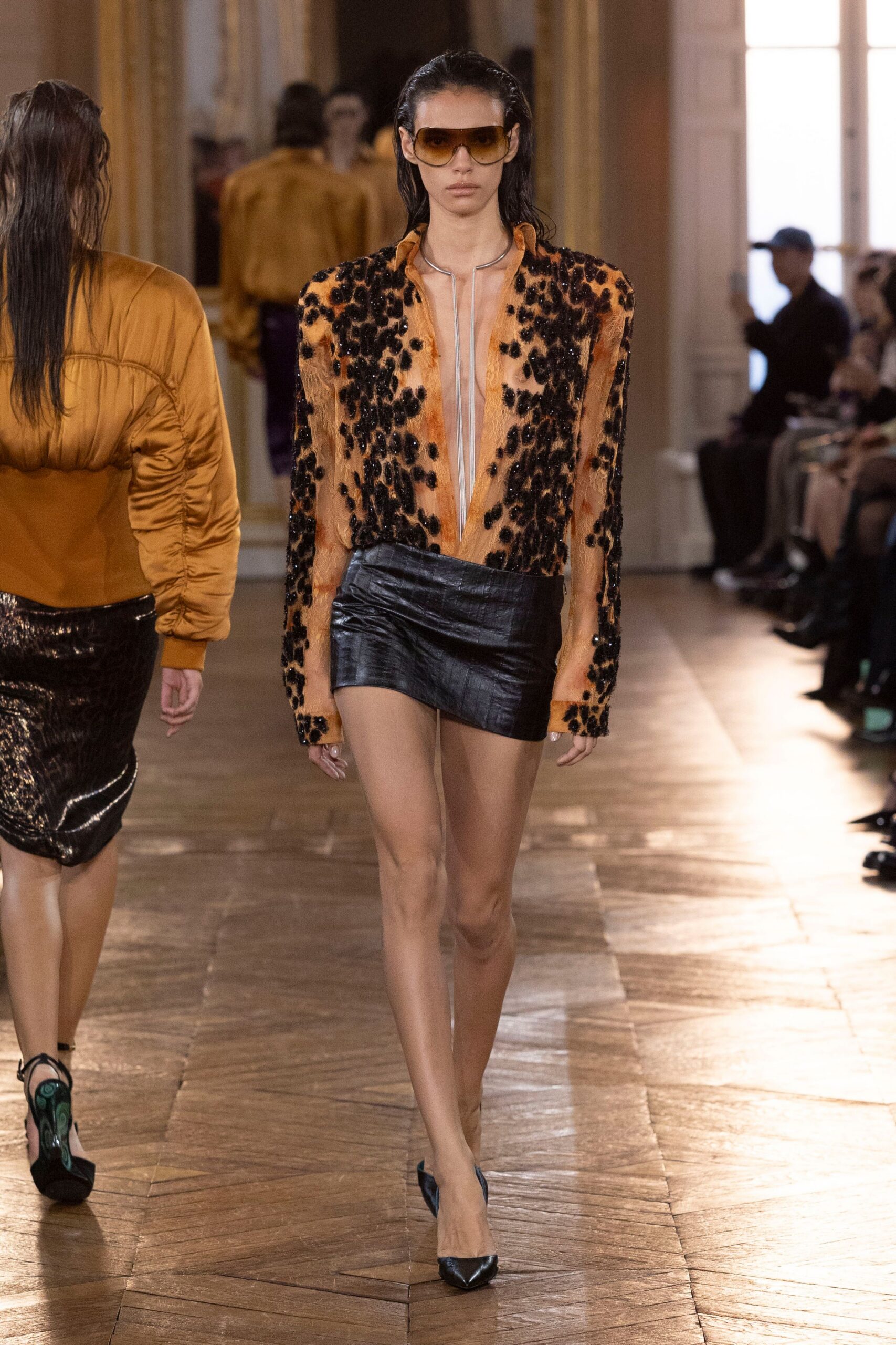 Amber aviators with a broad gradient lens. Peach sheer bodysuit scattered with black sequins, plunged to the navel. Silver collar dropping in a narrow U down the chest. Black leather miniskirt wrapped low, one corner kicked upward. Black pointed pumps. Crossing a hotel ballroom floor before the auction starts.