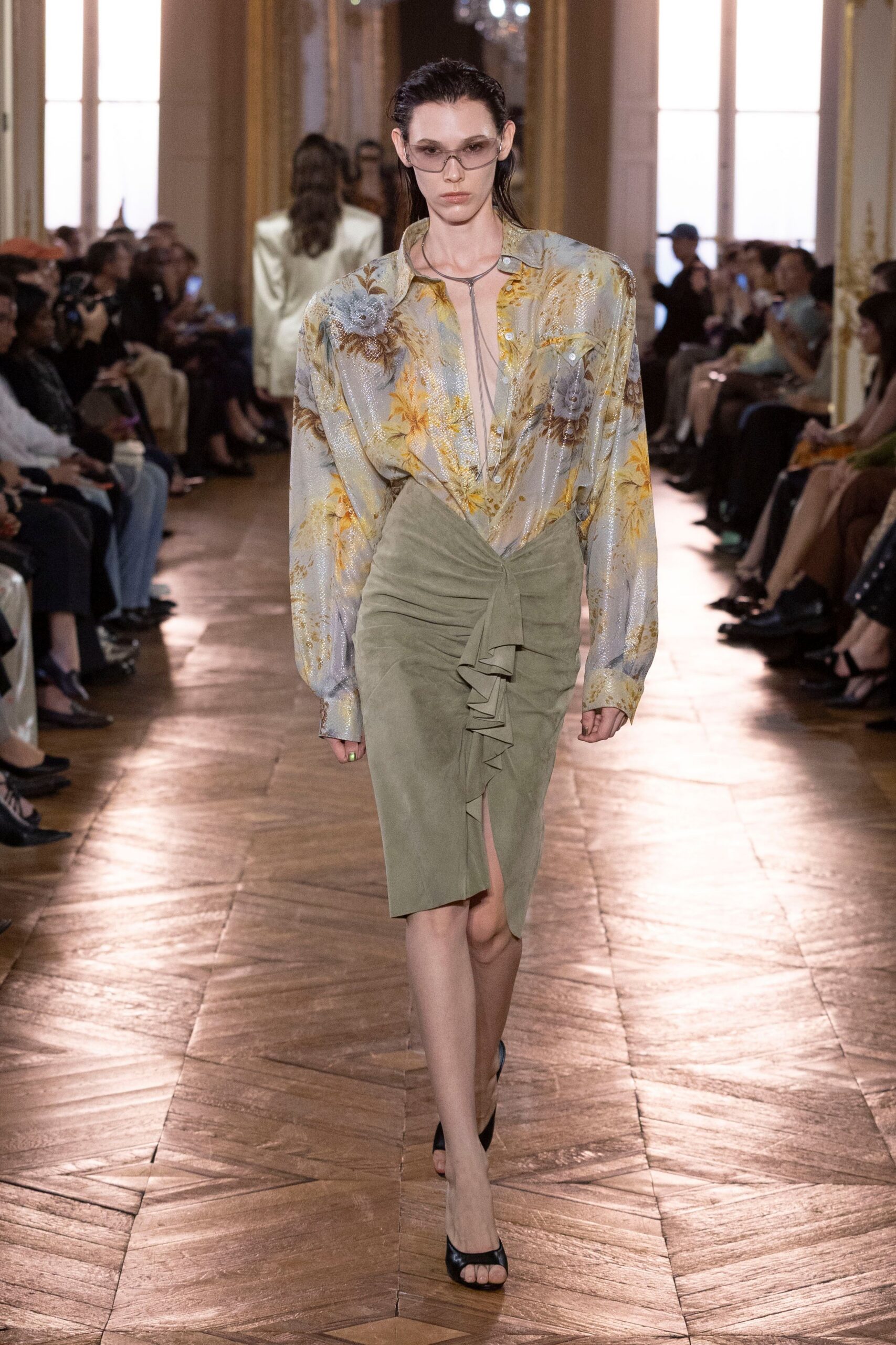 Smoky wraparound sunglasses with a narrow silver bridge. Shimmering pale floral shirt, worn open to a deep plunging V. Long silver lariat dropping straight down the bare chest. Sage suede wrap skirt, ruched tight with a ruffled front tie. Black open-toe mules. Hotel lobby after rain, cutting across to steal five quiet minutes.