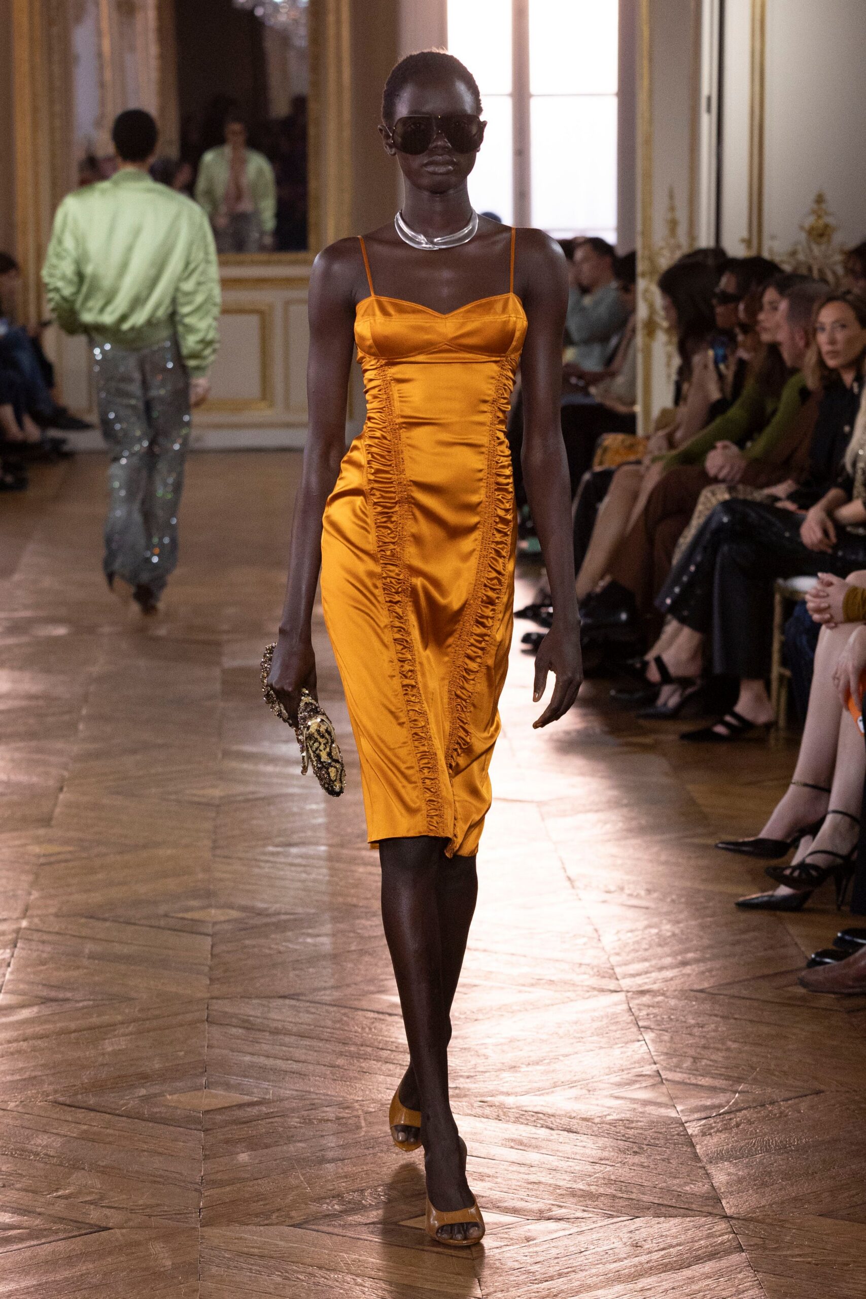 Oversized square sunglasses with a warm brown tint. Thick silver collar sitting open at the throat. Saffron satin slip dress, ruching laddered down both side seams. Beaded python clutch with a dangling chain handle. Ochre peep-toe mules. Crossing a hotel ballroom before the wedding band starts.