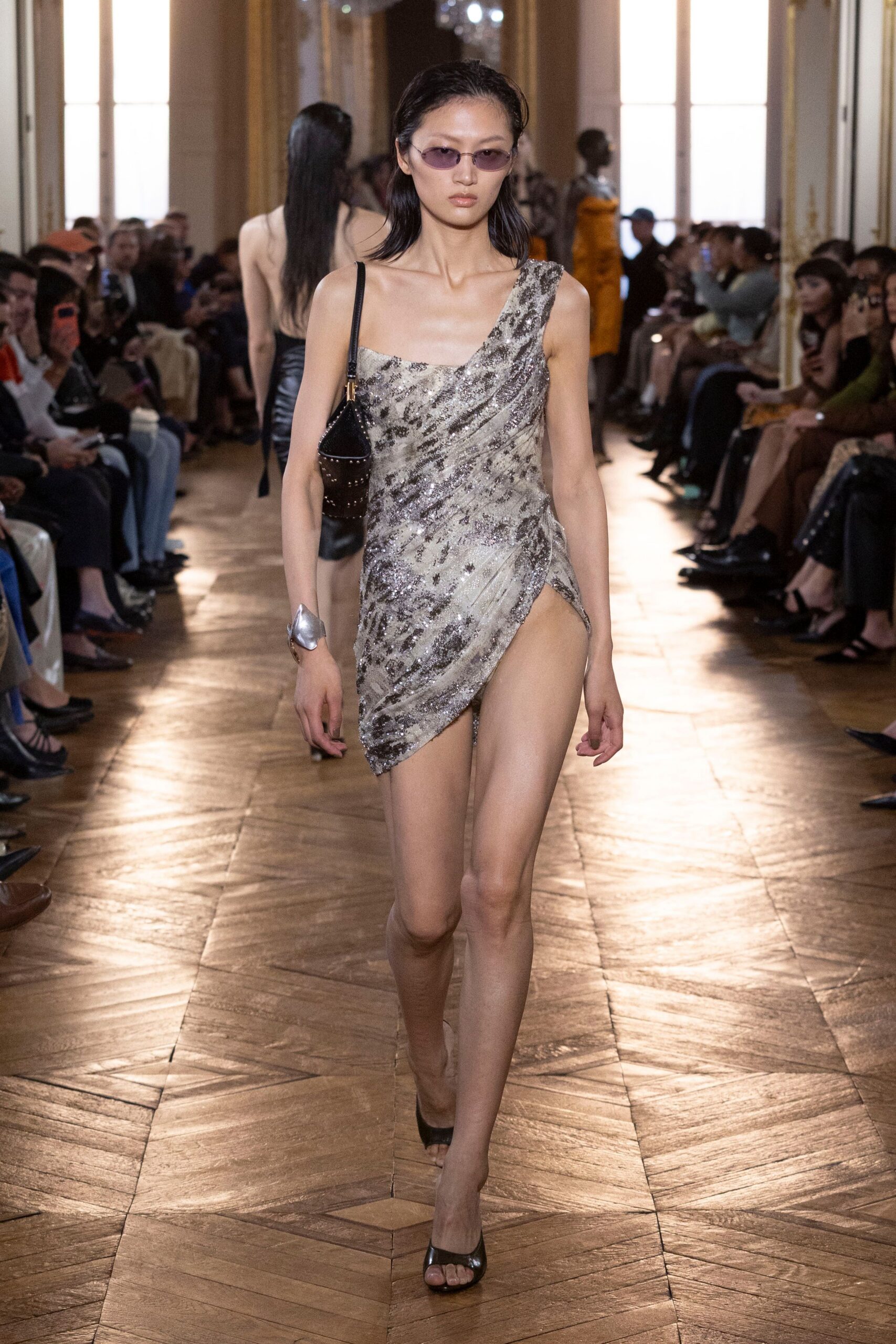 Narrow mauve-tinted lenses in a slim silver wire frame. Asymmetric silver sequined mini, ruched tight with a thigh-high wrap slit. Black studded shoulder bag tucked under one arm. Molten silver cuff, petal-shaped at the wrist. Black peep-toe mules. Crossing a hotel ballroom after midnight, still glitter on the parquet.