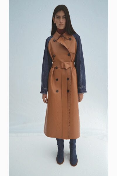 Camel double-breasted coat with indigo denim sleeves and laser-cut edge trim. Burgundy knit turtleneck rising close under the collar. Self belt with a broad rectangular buckle at the waist. Blue suede knee boots, toes rounding softly from the hem. Cold morning outside a civic building, papers folded under one arm.